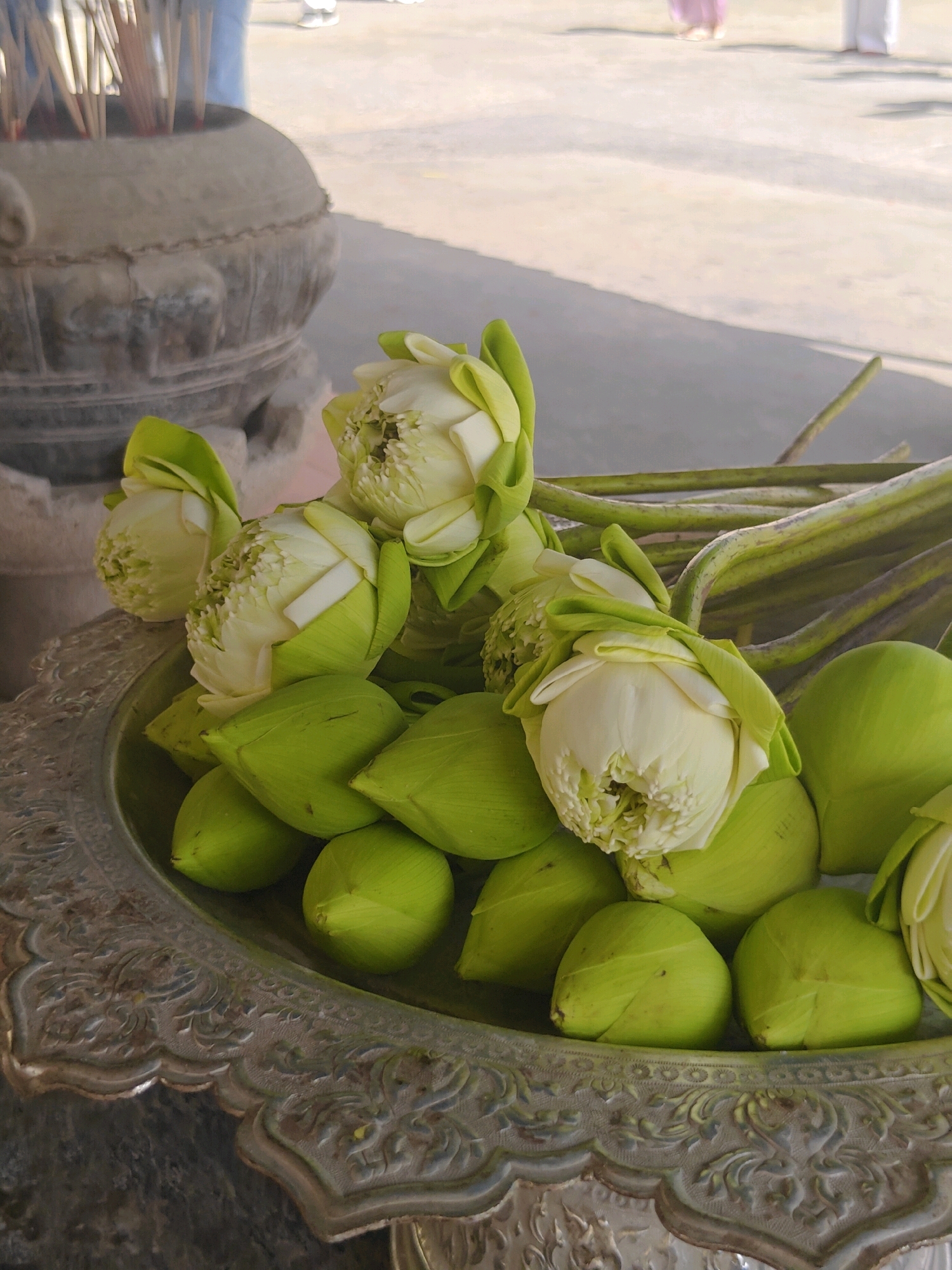 A close-up, eye-level shot depicts a silver ornate bowl filled with a collection of unopened and partially opened white lotuses with vibrant green sepals. The lotuses are arranged densely, with their stems extending upwards and to the right. In the background, a stone incense burner, filled with numerous incense sticks, is visible. The ground appears to be a paved surface, possibly outdoors. In the far background, blurred figures, likely people, are present, one wearing a pink garment and another in white. The lighting suggests a daytime setting, with some shadows cast on the ground. The scene is set in Phra Nakhon Si Ayutthaya City Municipality, Thailand, likely at a temple or cultural site.