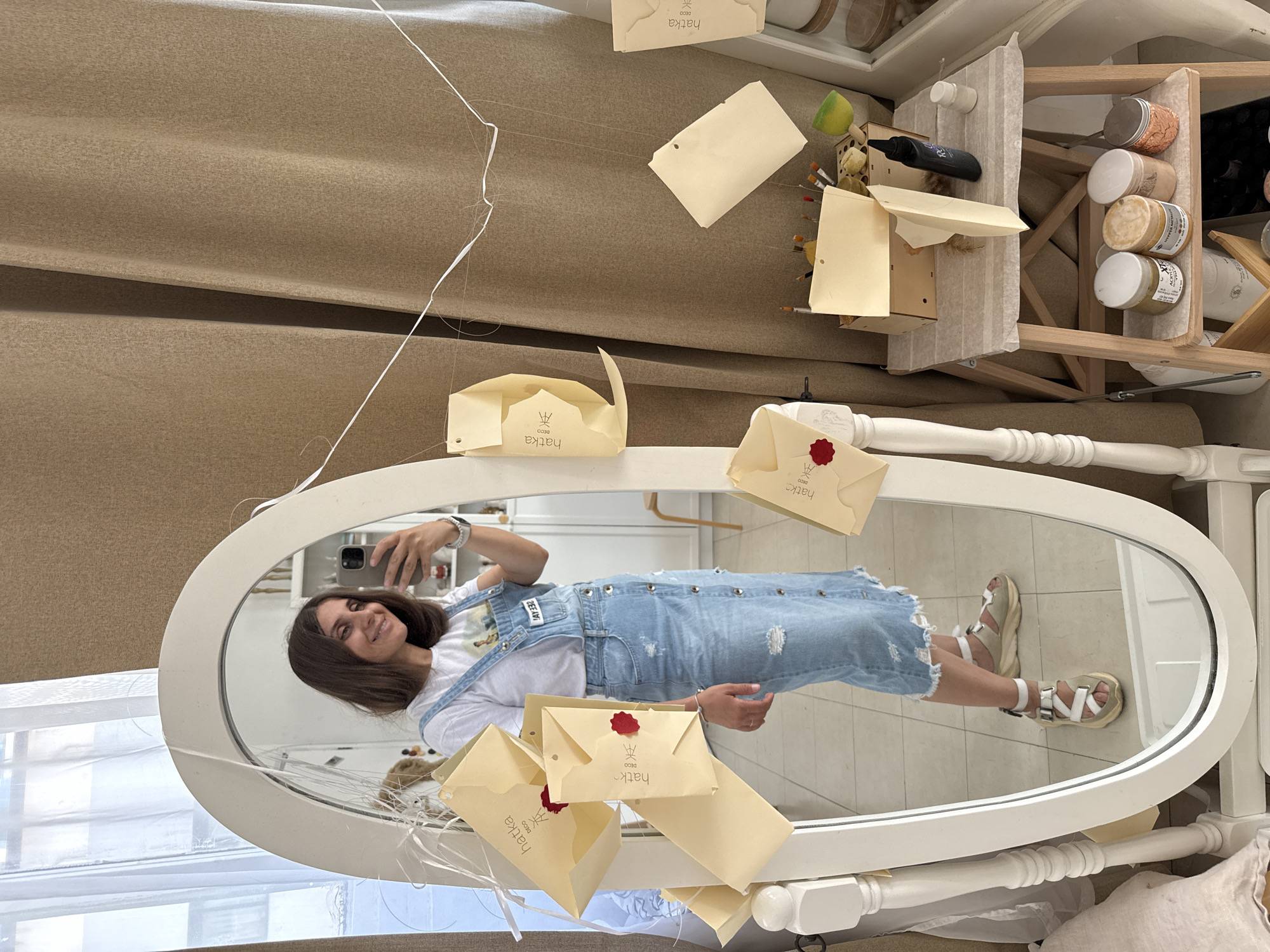 A young woman is taking a selfie in a mirror in what appears to be a studio space. The woman is wearing a white t-shirt with a graphic print and a denim overall dress. She has medium-length brown hair and is smiling at her phone, which she is holding in her right hand. The mirror is large and oval-shaped with a white wooden frame. It is leaning against a wall covered with textured beige curtains. White strings are suspended from the ceiling, holding up several yellow envelopes with red wax seals and the logo "hatka deco". Some of these envelopes are also reflected in the mirror. To the right of the mirror, a small wooden step stool is visible, decorated with more yellow envelopes and various craft supplies, including jars of paint or pigment, brushes, and a bottle of glue. The floor is tiled. The scene suggests a creative or artistic environment. The presence of the logo "hatka deco" could potentially place the location in Tel Aviv, Israel, as "Hatka" is a common surname and business name in Israel.