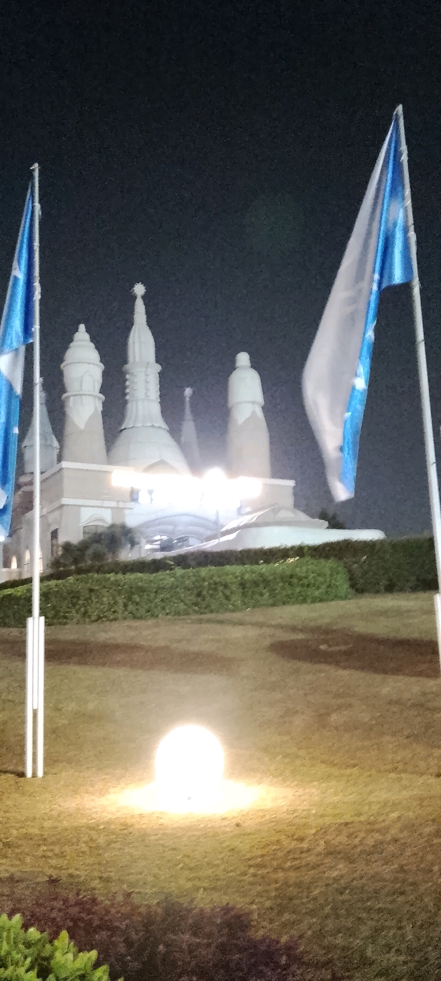 A night-time, medium shot captures a building with multiple spires and domes, illuminated by bright lights. Two flags, one on the left and one on the right, are positioned in the foreground, appearing to be on flagpoles. The flag on the left is mostly visible, while the flag on the right is partially obscured. In the middle ground, a well-maintained hedge runs horizontally across the image, separating the lawn from the building. A spherical light fixture sits on the lawn in the foreground, emitting a bright glow. The background is a dark, overcast sky. This image depicts the front of the building complex of the Sri Sathya Sai Baba Center in Gunung Putri, Indonesia.