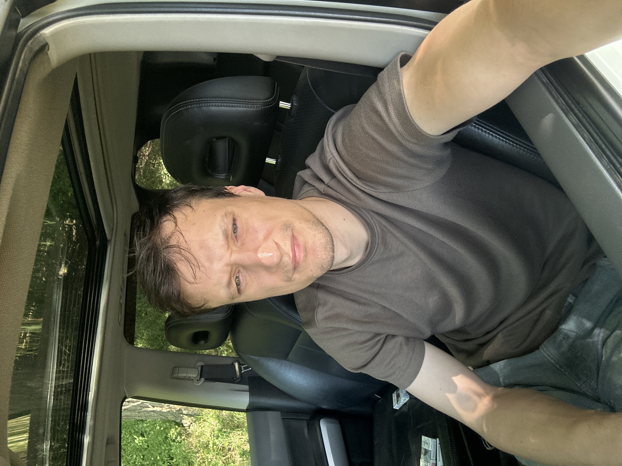 A man is sitting in the driver's seat of a car with a sunroof open. He is looking towards the camera and has a serious expression on his face. He is wearing a brown t-shirt and gray pants. The interior of the car is visible, including black leather seats. Outside the car, there are trees and greenery. There is a pack of cigarettes on the seat next to him. The image appears to have been taken in Makiv, Ukraine, based on the context provided in the prompt.