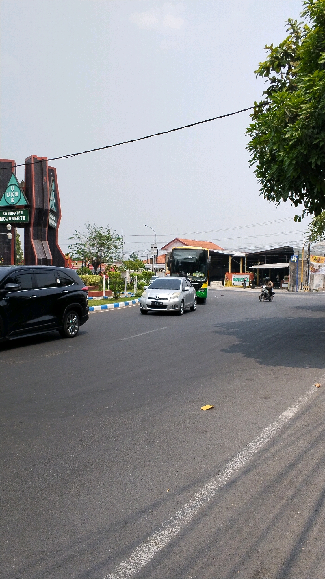 The image captures a street scene in Banjaragung, Indonesia, likely near a landmark or entrance to the regency. In the foreground, a black SUV is parked on the left side of the asphalt road. To the right of the SUV, a silver sedan is driving in the same direction. Further down the road, a yellow and green bus is in motion, and a person on a motorcycle is visible to the right of the bus. Behind the vehicles, there are buildings and lush green trees. A prominent triangular monument with "UKS" and "KABUPATEN MOJOKERTO" inscribed on it stands on the left side of the frame, indicating this as the entrance to Mojokerto Regency. The sky is overcast, suggesting a bright but possibly hazy day. The road is marked with white lines, and shadows from trees stretch across the pavement, indicating the sun is positioned somewhere to the right.