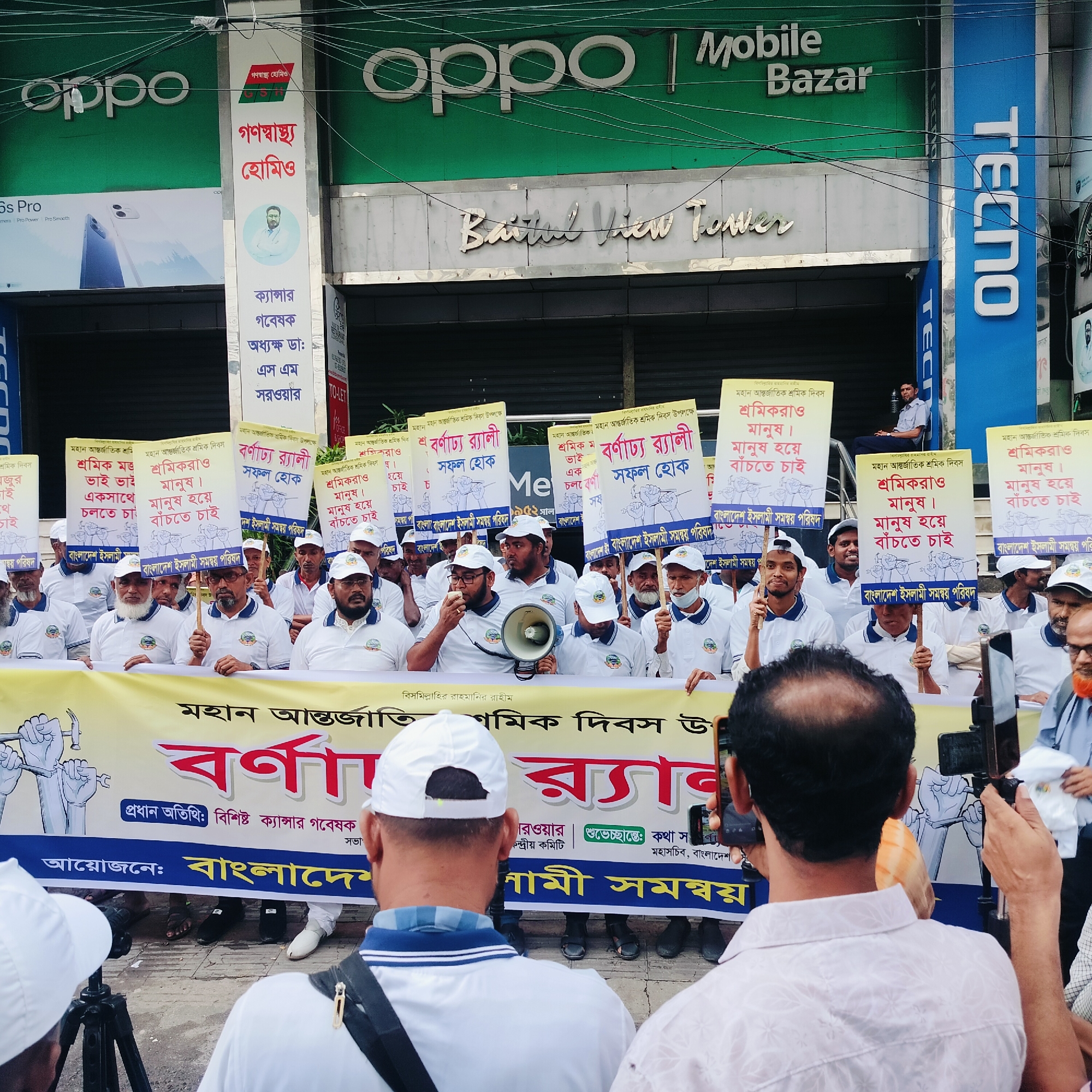 A daytime scene captures an outdoor rally or demonstration taking place on a street in Dhaka, Bangladesh. A large group of men, mostly middle-aged to elderly, are gathered, uniformly dressed in white polo shirts with a logo and white caps. In the immediate foreground, two individuals are seen from behind, holding up smartphones to record or photograph the event.
The participants hold numerous white placards featuring Bengali text in red and black, and a prominent yellow banner spans the width of the gathering. A man in the center of the crowd is speaking into a white megaphone. The placards commemorate "মহান আন্তর্জাতিক শ্রমিক দিবস উপলক্ষে" (On the occasion of Great International Workers' Day) and convey messages such as "শ্রমিকরাও মানুষ। মানুষ হয়ে বাঁচতে চাই।" (Workers are also humans. We want to live as humans.) and "বর্ণাঢ্য র্যালী সফল হোক।" (May the colorful rally be successful.). The organizing body, "বাংলাদেশ ইসলামী সমন্বয় পরিষদ" (Bangladesh Islamic Coordination Council/Committee), is identified on the placards. The large yellow banner repeats "বিসমিল্লাহির রাহমানির রাহীম" (In the name of Allah, the Most Gracious, the Most Merciful), mentions the "মহান আন্তর্জাতিক শ্রমিক দিবস" (Great International Workers' Day), and highlights the "বর্ণাঢ্য র্যালী" (Colorful Rally). It also lists the "প্রধান অতিথি: বিশিষ্ট ক্যান্সার গবেষক এস এম সরওয়ার" (Chief Guest: Distinguished Cancer Researcher S M Sarwar).
The backdrop consists of commercial buildings. Visible signs include "Oppo," "Mobile Bazar," "TECNO," and "Baitul View Tower." A vertical sign for "গণস্বাস্থ্য হোমিও" (Ganashasthya Homoeo) features a portrait and identifies "ক্যান্সার গবেষক অধ্যক্ষ ডা: এস এম সরওয়ার" (Cancer Researcher Principal Dr: S M Sarwar), linking to the chief guest mentioned on the banner. The sky appears overcast, providing soft, diffused lighting. Overhead utility wires crisscross the upper part of the image. The mood of the gathering appears to be one of organized protest or advocacy.