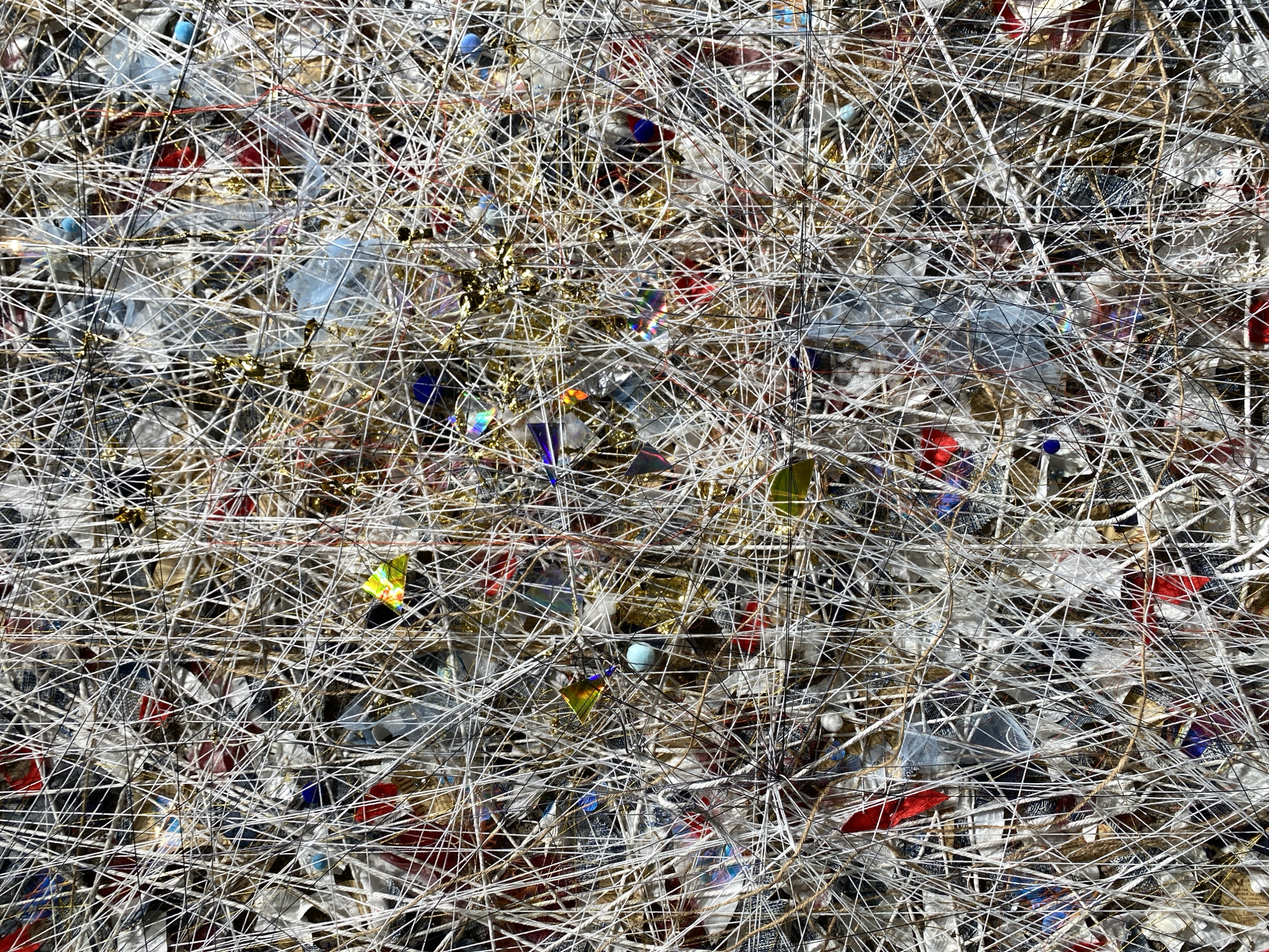 This is a close-up, abstract, still life photograph of a densely woven tapestry of tangled white and dark threads, interspersed with various small objects. These objects include iridescent geometric shards, small blue beads, scraps of red and blue fabric, and shiny gold glitter. The overall impression is chaotic and textured, with the threads creating a complex grid that obscures and reveals the embedded items. The image does not depict any specific location in Yuzhong District, China, nor does it contain any people, actions, or interactions. It is a purely visual composition focused on texture, color, and form.