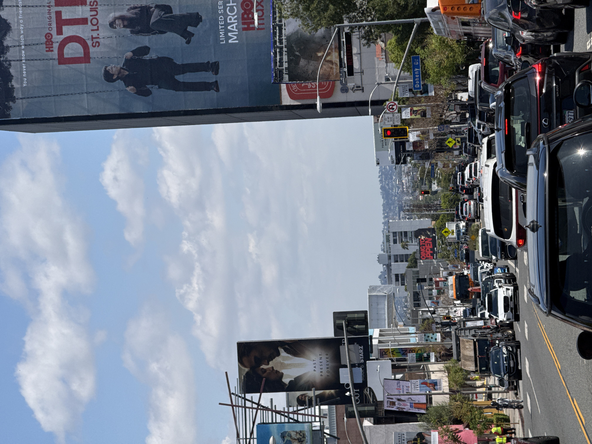 The image shows traffic congestion on a city street with numerous billboards on display. The billboards advertise various shows and movies. Street signs indicate the location is at the intersection of Sunset Strip and Doheny Drive in West Hollywood, California. The sky is partly cloudy, and the scene suggests a busy urban environment.