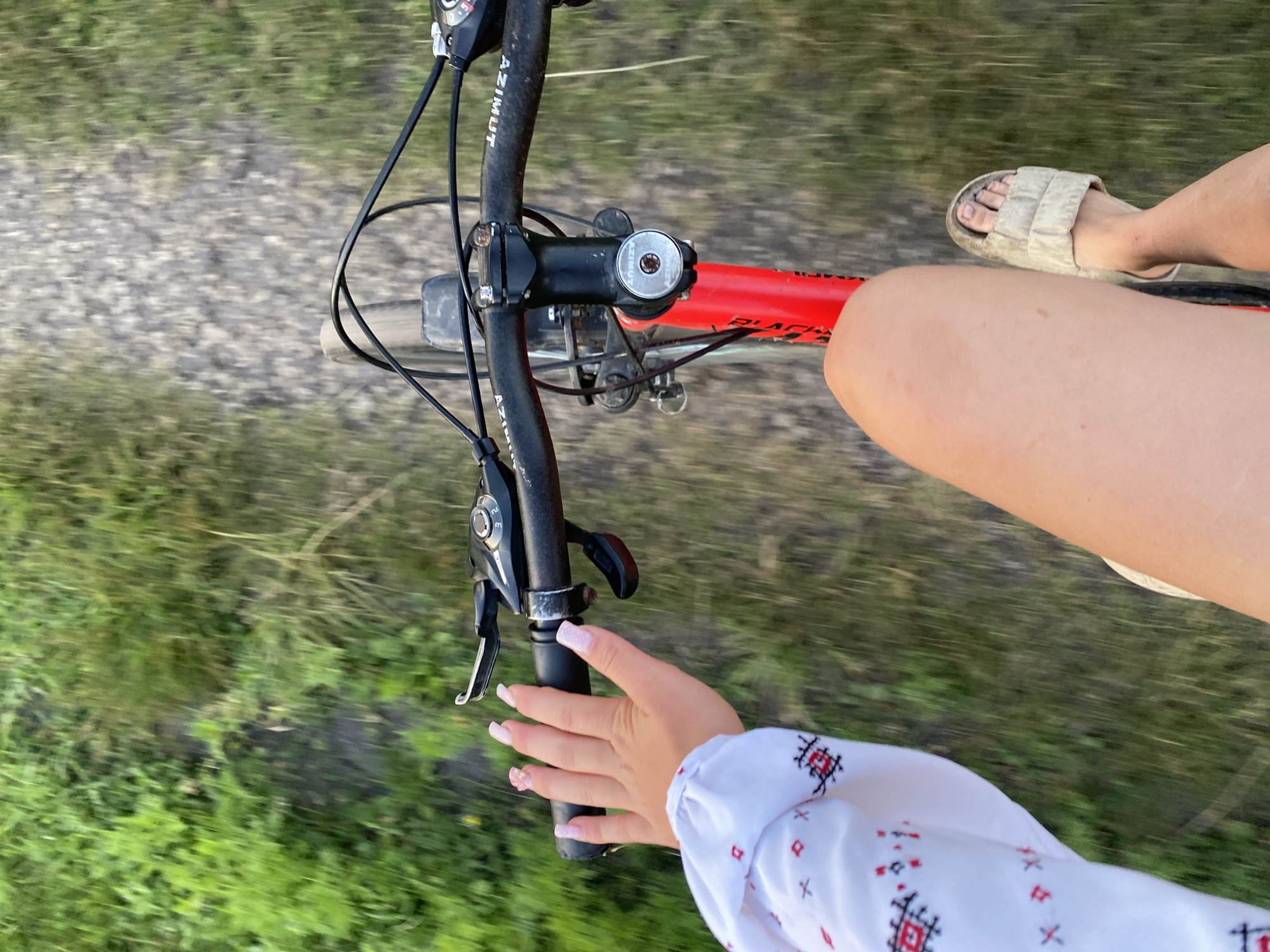 The image shows a first-person perspective of someone riding a bicycle. The subjects are the rider's legs and hand, the bicycle's handlebars, and the surrounding environment. The rider's left leg is visible, clad in a white sandal. The rider's right leg is also visible, bare and closer to the foreground. The rider's left hand, with manicured pink nails and small floral nail art, is gripping the left handlebar.
The bicycle's handlebars are black, with "AZIMUT" visible on them. A red bicycle frame is also visible. The scene is outdoors, with a path of gravel or dirt and green grass or foliage on either side, suggesting a natural or rural setting. The motion blur indicates the bicycle is in motion.
The time of day appears to be daytime, possibly late afternoon or early evening, judging by the lighting. The weather seems pleasant and suitable for outdoor activity. There are no strong emotional cues, but the activity of cycling suggests enjoyment or leisure. The setting is unclear, but the natural elements point to a location outside of a city.
Visible text includes "AZIMUT" on the handlebars.