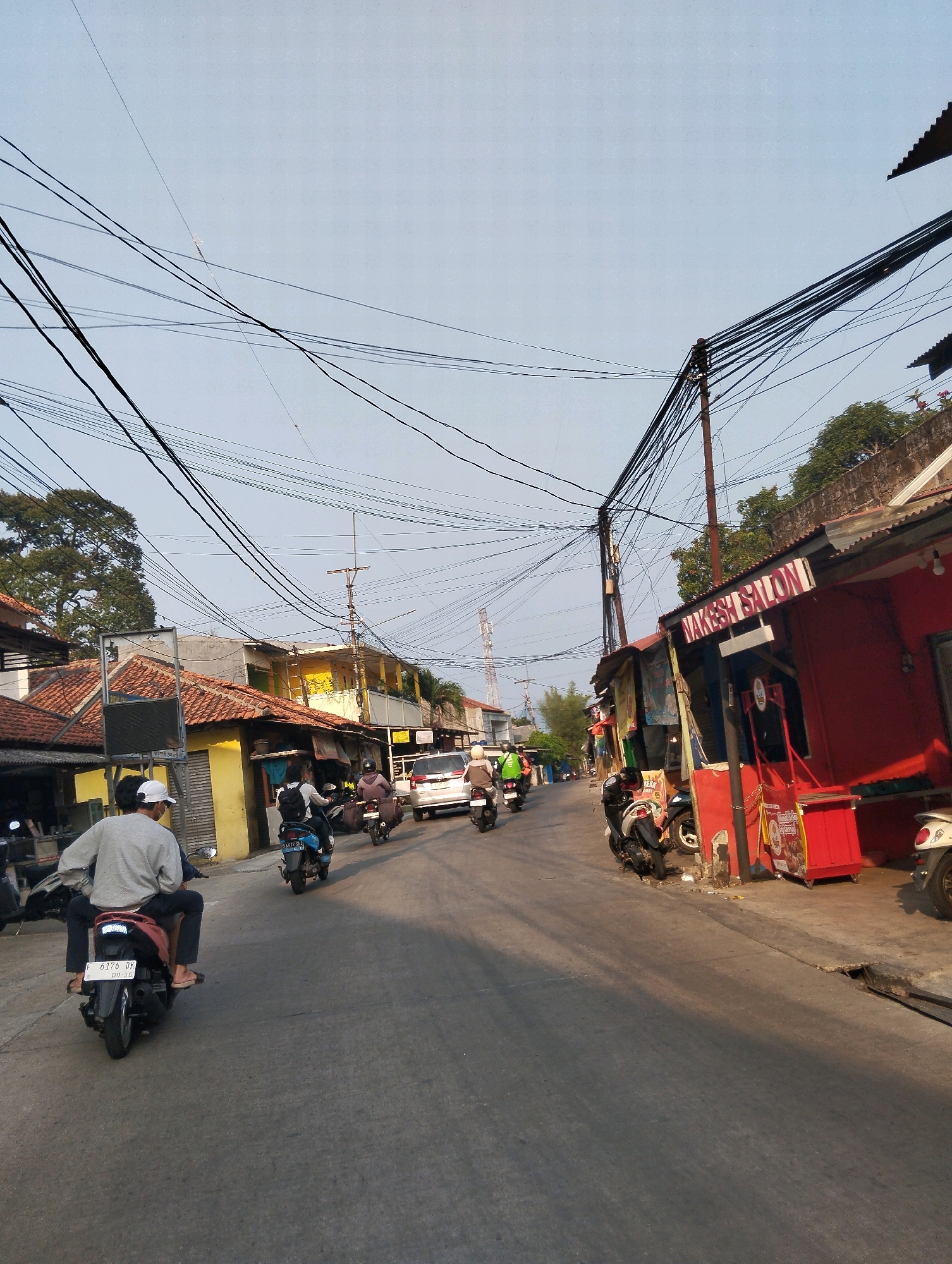 The image depicts a street scene in Sukaraja, Indonesia, characterized by an abundance of overhead power lines. The road surface is asphalt, with a gutter present on the right side. A group of motorcycles and one car are visible, suggesting moderate traffic.
On the left side of the street, there are buildings with tiled roofs, some painted yellow and others with a mix of colors. One building on the left appears to be a shop with a closed shutter. A person wearing a gray sweatshirt and a white cap is riding a motorcycle in the foreground, with another person seated behind them.
On the right side of the street, there is a red building with a sign that reads "NAKESH SALON". Other commercial establishments with awnings and signs are also present. Several motorcycles are parked along the sidewalk. Trees and vegetation are visible behind some of the buildings. The sky is clear and pale blue, indicating daylight. The lighting suggests it might be late afternoon or early morning due to the long shadows cast on the road.