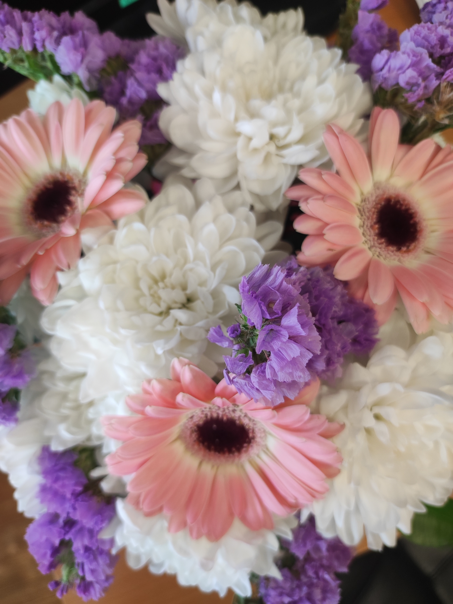 The image shows a bouquet of flowers. There are white chrysanthemums, pink gerbera daisies, and purple statice flowers. The flowers are arranged in a cluster, with the white chrysanthemums making up the bulk of the arrangement. The pink gerberas are interspersed throughout, and the purple statice adds pops of color. The arrangement appears to be in a vase or container, though this is not fully visible in the image. The location in the city of Votkinsk, Russia is not evident from the image. There are no visible people, subjects, or actions depicted in this image.