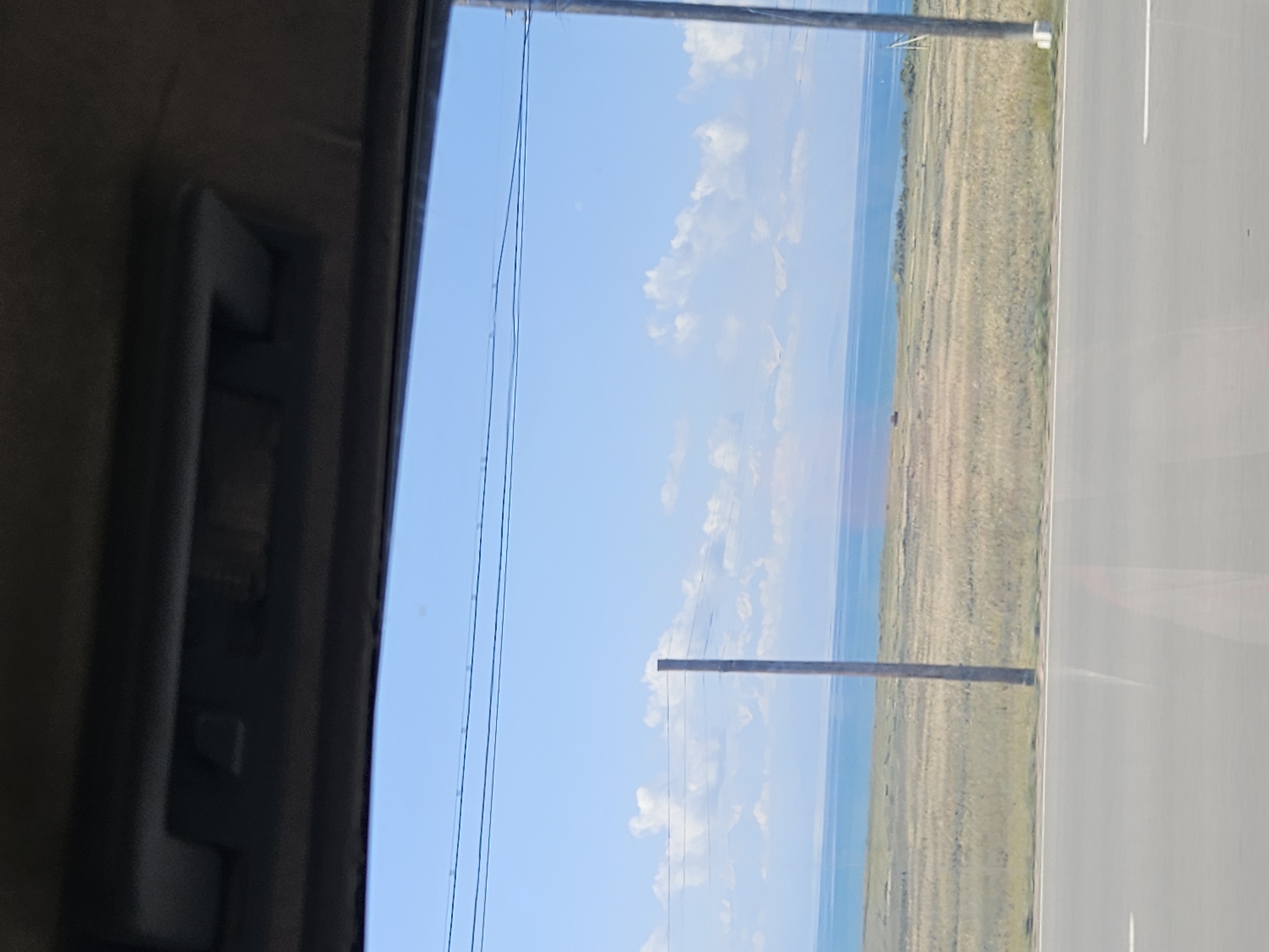 The image shows a view from inside a vehicle looking out a window. The window frame is dark and partially visible at the top of the image. Through the window, a wide expanse of a body of water, possibly a lake or sea, is visible under a bright blue sky with scattered white clouds. The water appears calm and a light blue color.
In the foreground, a flat, dry, grassy plain stretches from the water's edge towards the road. The grass appears to be a mix of green and golden hues, suggesting it is dry or dormant. A few scattered, thin wooden poles, likely part of a fence or power line, are visible in the middle ground. One pole is particularly prominent in the center-left of the image, and another appears on the far right. In the distance, along the horizon where the water meets the sky, a faint line of hills or mountains can be discerned.
In the immediate foreground, a grey asphalt road is visible, with a white line marking its edge. Reflections of the sky and surrounding environment are visible on the road's surface. The overall scene depicts a natural, open landscape under a clear sky. There are no people or specific landmarks identifiable in the image that would pinpoint its exact location, but it captures a serene, rural or natural setting.