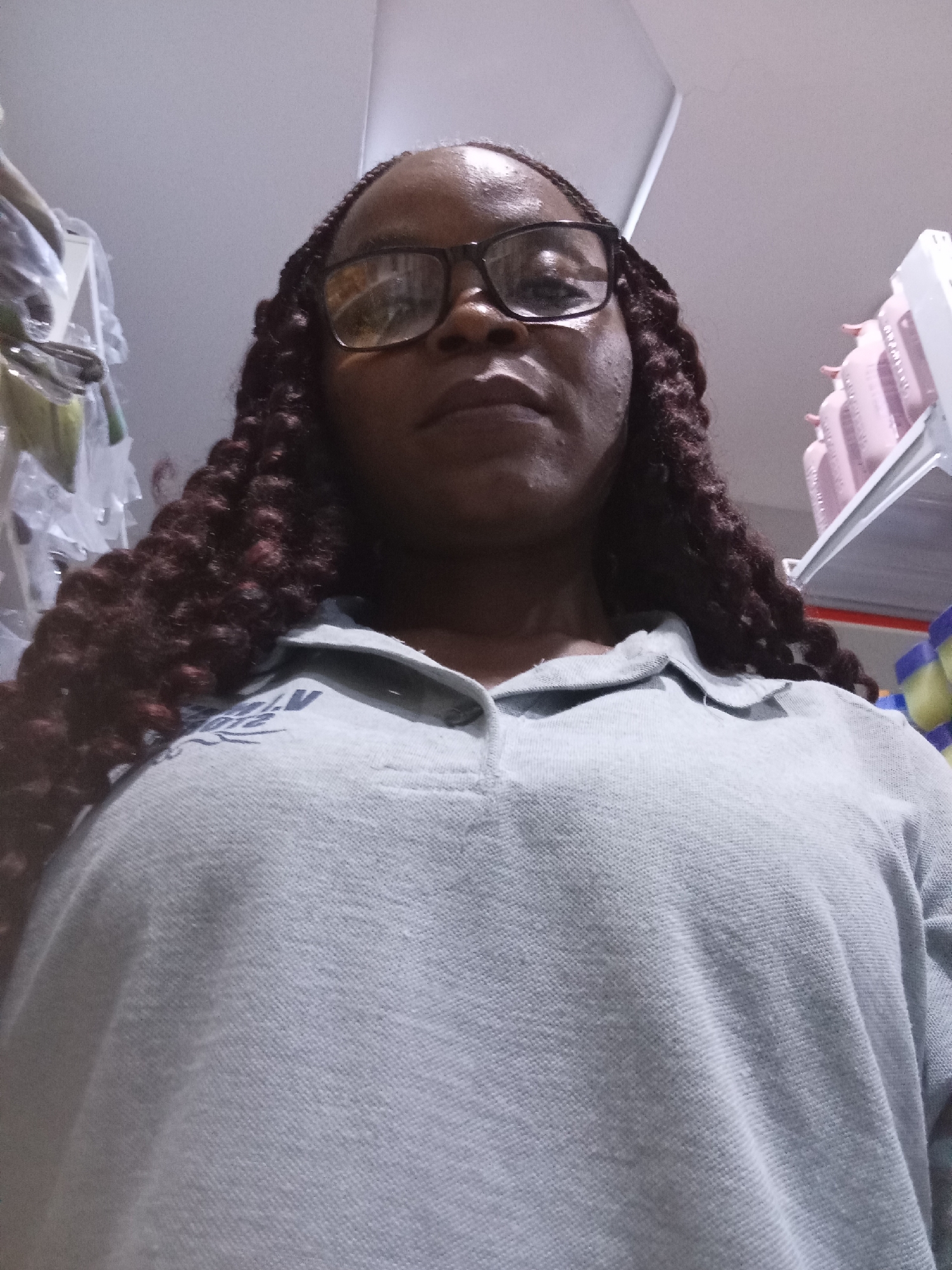 A low-angle, close-up shot focuses on a Black woman from the chest up, with her head tilted back and looking directly at the camera. She is wearing glasses with thick black frames and has dark brown braided hair that falls down her shoulders. Her skin is dark and appears slightly oily. She is wearing a light blue polo shirt with a collar and a logo on the left chest that is partially visible, appearing to be a dark emblem with white lettering.
The background consists of shelves stocked with various items. On the left, there are clothes hanging in clear plastic packaging. To the right, there are rows of pink bottles on a white shelf, and further to the right, there are yellow and blue cylindrical objects. The lighting suggests an indoor environment, possibly a retail store. The overall scene is a personal photograph, likely a selfie, taken in a commercial setting. There is no visible indication that this location is specifically in Howayi, Nigeria.