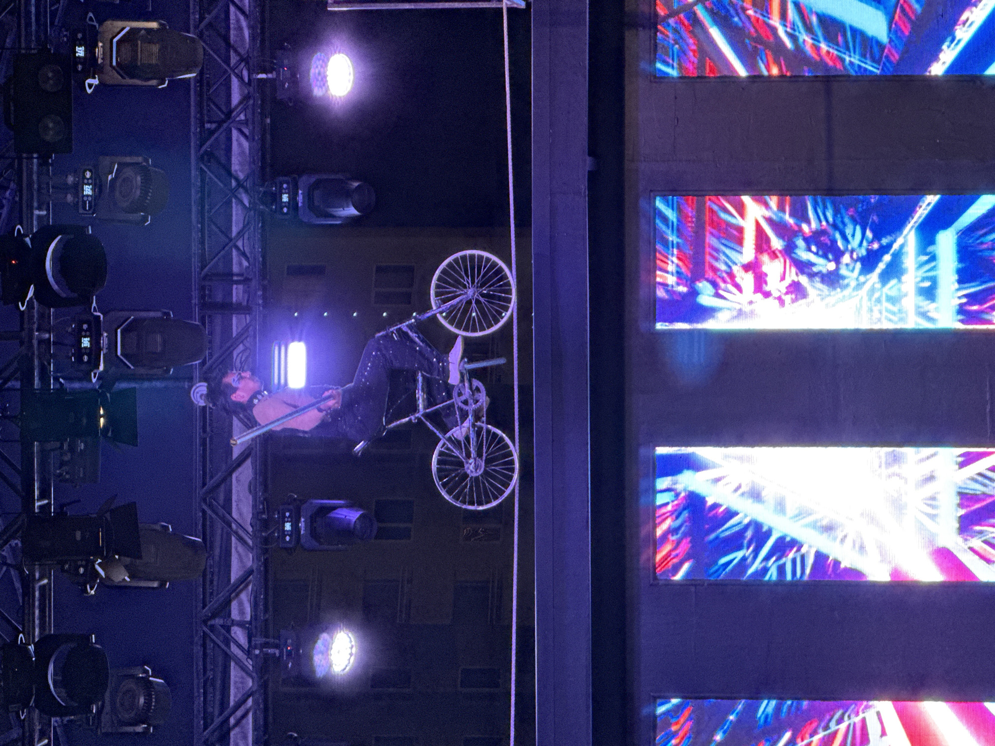 A person balances on a tightrope while riding a small bicycle. The person is wearing a dark, sequined outfit and a choker necklace, with their hair styled in a bun. They are holding a long pole horizontally for balance. The tightrope is suspended above a stage area, with numerous lights hanging from above. Below the stage are large vertical screens displaying vibrant, abstract light patterns in red, blue, and white. The background appears to be a dark building with illuminated windows. The scene suggests a performance or entertainment event. The location is Aksu, Turkey.