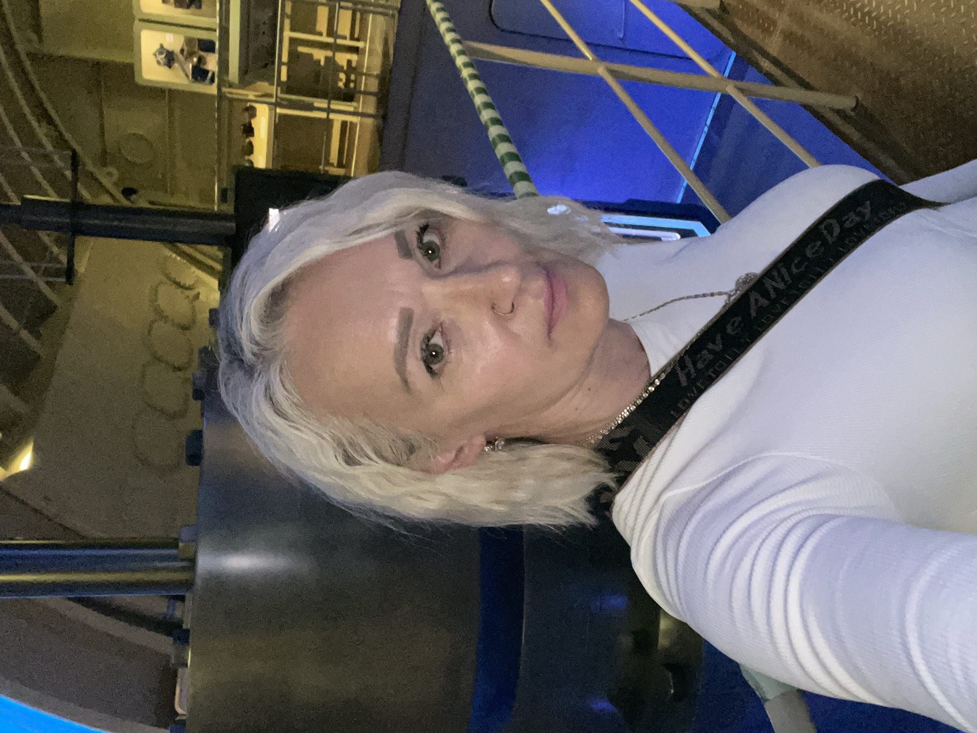 In Kronstadt, Russia, a woman with short, wavy, platinum blonde hair takes a selfie in what appears to be a museum or exhibition space. She is wearing a white, long-sleeved, ribbed shirt and a black strap with white text that reads "Have A Nice Day". A silver chain with a pendant is visible around her neck. She has a nose ring and wears small earrings. The background is industrial, with large, dark cylindrical structures and metallic railings, some of which are painted with green and white stripes. Blue lighting illuminates the background, suggesting an artificial environment. The woman is positioned in the foreground, looking directly at the camera with a neutral expression.