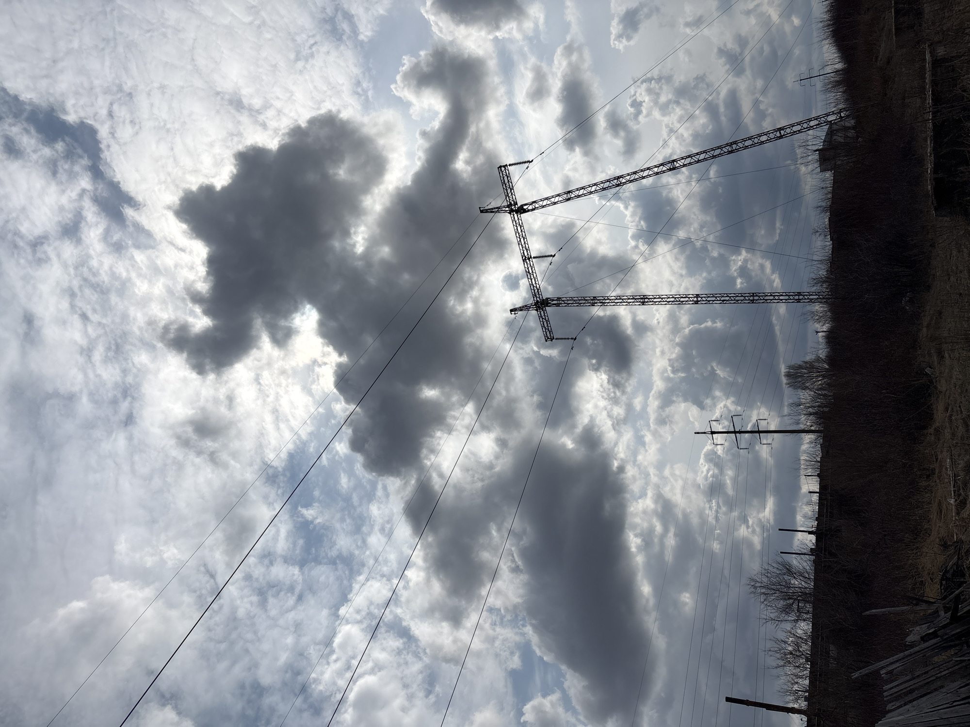 The image shows a view upwards towards the sky. Overcast with clouds, and a break in the clouds revealing blue sky. Power lines and power line towers are present. On the right side of the frame is an area with trees and foliage. Although not explicitly stated to be located in Ulyanovsk, Russia, the description is based solely on the content of the image. The image does not contain information about people, subjects, objects, actions, or interactions.