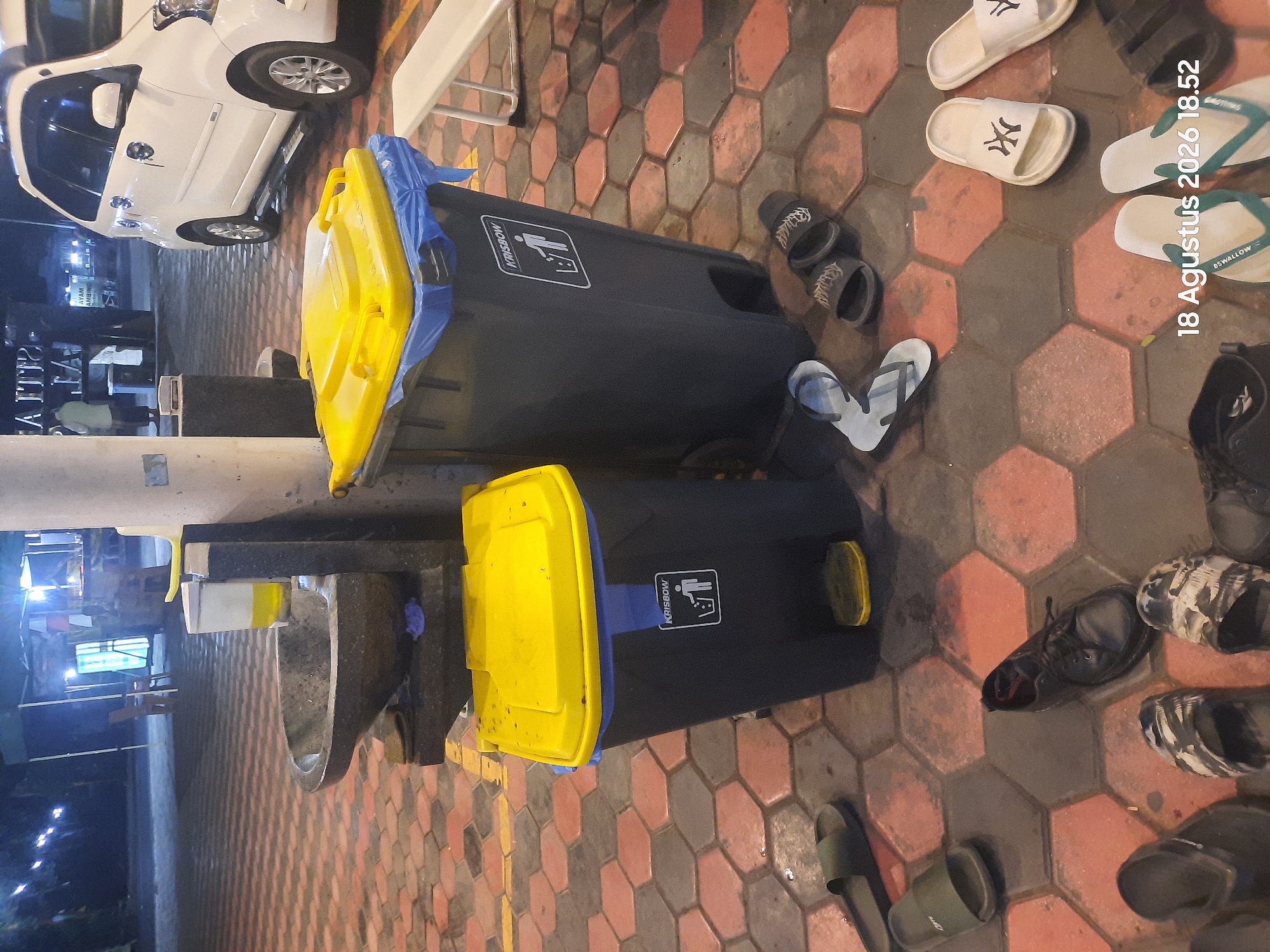In Sukokerto, Indonesia, at night, a scene unfolds at what appears to be an outdoor eatery or rest stop. Two dark grey trash bins with bright yellow lids stand prominently in the foreground, one with a blue liner visible. The bin on the right displays a white "KRISBOW" logo with a symbol of a person discarding waste. Nearby, a stone basin with a dispenser holding yellow liquid sits atop a wooden structure.
The ground is paved with hexagonal tiles in shades of reddish-brown and grey. Several pairs of footwear are scattered across the tiles, including dark green slides, black flip-flops, white flip-flops with a New York Yankees logo, and black lace-up shoes. In the background, a white minivan is parked, and indistinct figures can be seen near a tented area with bright lights, suggesting a bustling atmosphere. A white bench is also visible to the right of the trash bins. A date stamp at the bottom right reads "18 Agustus 2026 18.52".