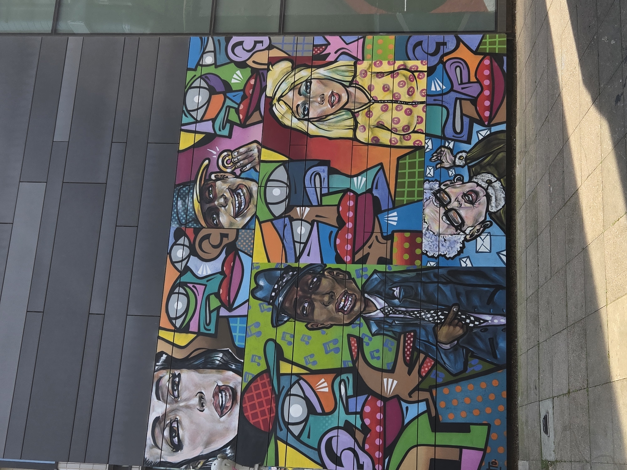 A colorful mural adorns a building facade, likely in London, United Kingdom. The artwork features portraits of well-known figures rendered in a bold, pop art style. The mural comprises individual blocks, with the faces seemingly split into distinct sections, adding a stylized and abstract effect. The overall environment suggests an urban setting, marked by the presence of buildings with modern architectural elements.