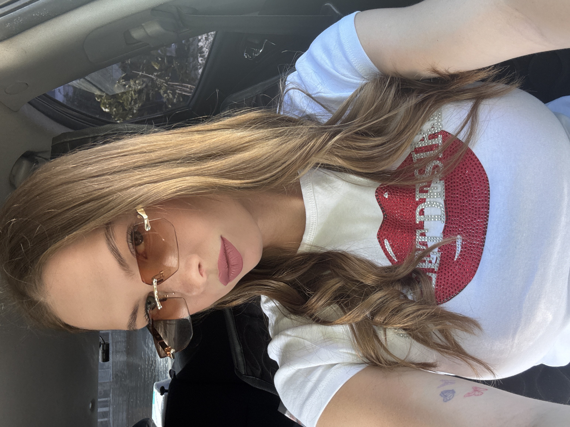 A young woman with long, brown hair and light skin is sitting in the driver's seat of a car. She is wearing sunglasses with amber lenses and a white t-shirt with a red sequined lip design. The background shows the interior of the car and a glimpse of the outside. The image does not provide any information to ascertain the location in the city of Perm, Russia.