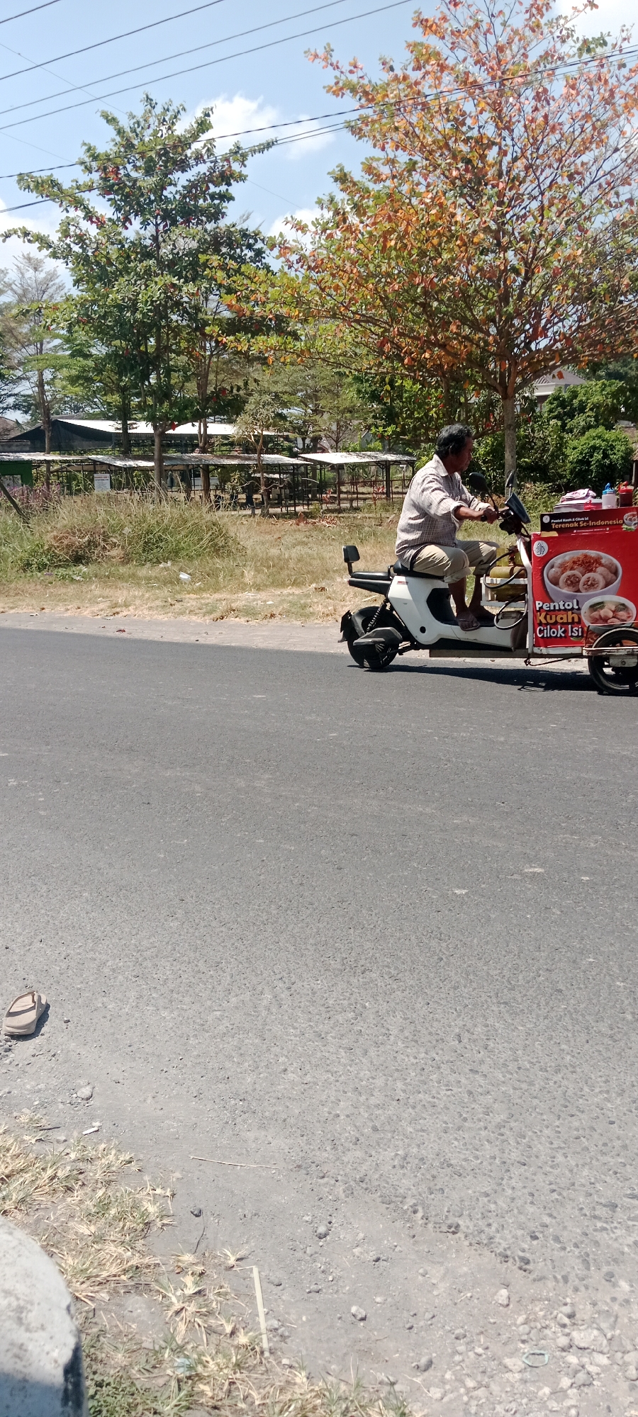 An elderly Indonesian man is riding a white electric scooter with a food cart attached. The scooter is moving on a paved road. The man is wearing a plaid shirt and light-colored pants. The food cart has a red sign with text and an image of food. The environment is outdoors with trees and a bright sky. In the background, there are some covered structures which appear to be stalls or outdoor seating areas. The ground on the side of the road is covered in dry grass and some litter. There is a single sandal on the road in the lower left corner of the image. The image appears to have been taken in Maguwoharjo, Indonesia, based on the provided context.