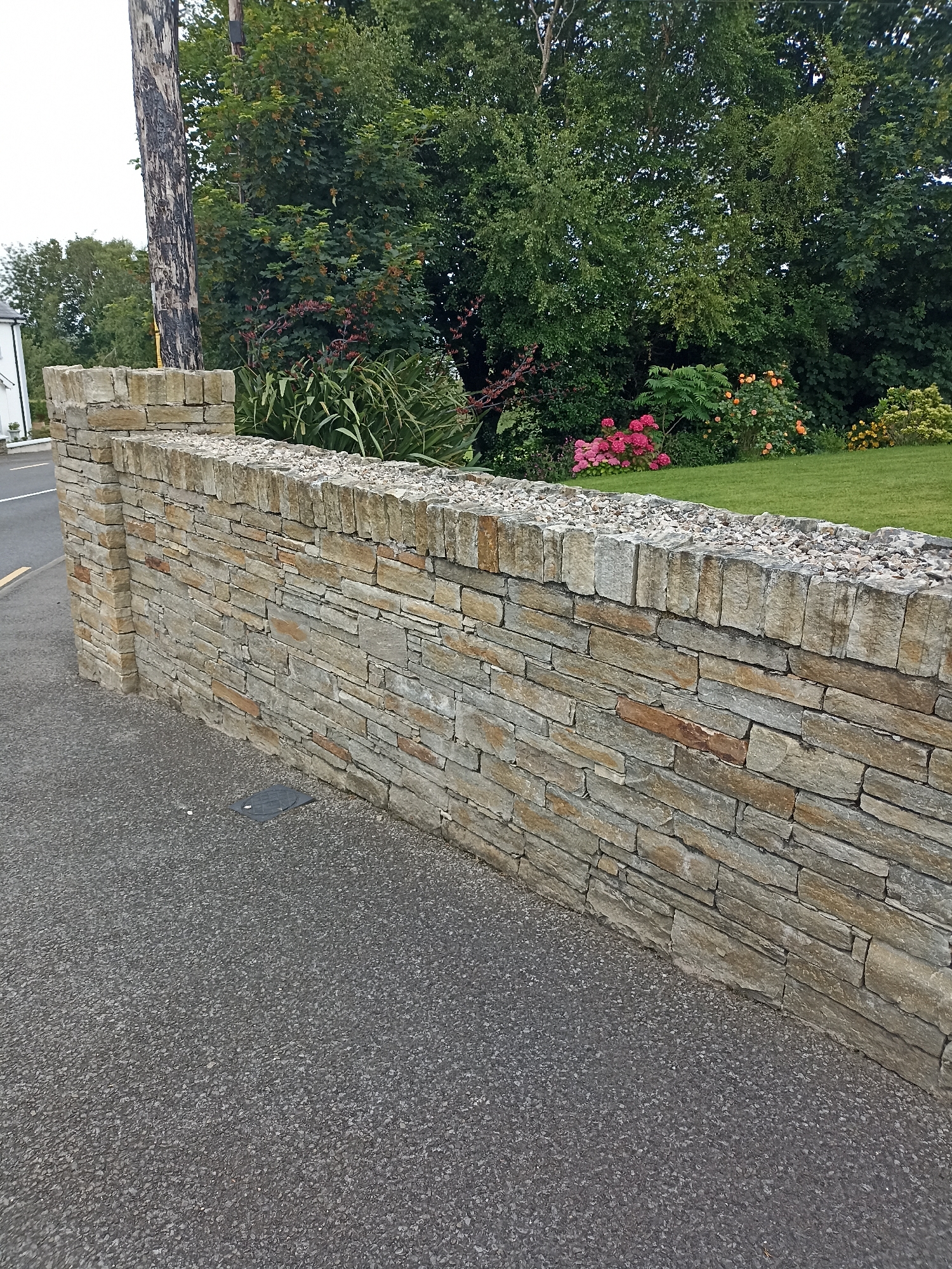 A stone wall runs along the side of an asphalt road. The wall is constructed from flat, rectangular stones of varying sizes and shades of brown and grey, laid horizontally. The top of the wall is capped with smaller stones and gravel. A square, dark-colored drain cover is visible on the asphalt next to the wall. Behind the wall, a lush green lawn is visible, bordered by a variety of trees and flowering bushes. The trees are tall and dense, with dark green foliage. The bushes include vibrant pink and orange flowers. A wooden utility pole stands to the left of the wall, partially obscured by foliage. A white building with a pitched roof is visible in the distance on the left. The sky is overcast. The image appears to have been taken in Annagry, Ireland.