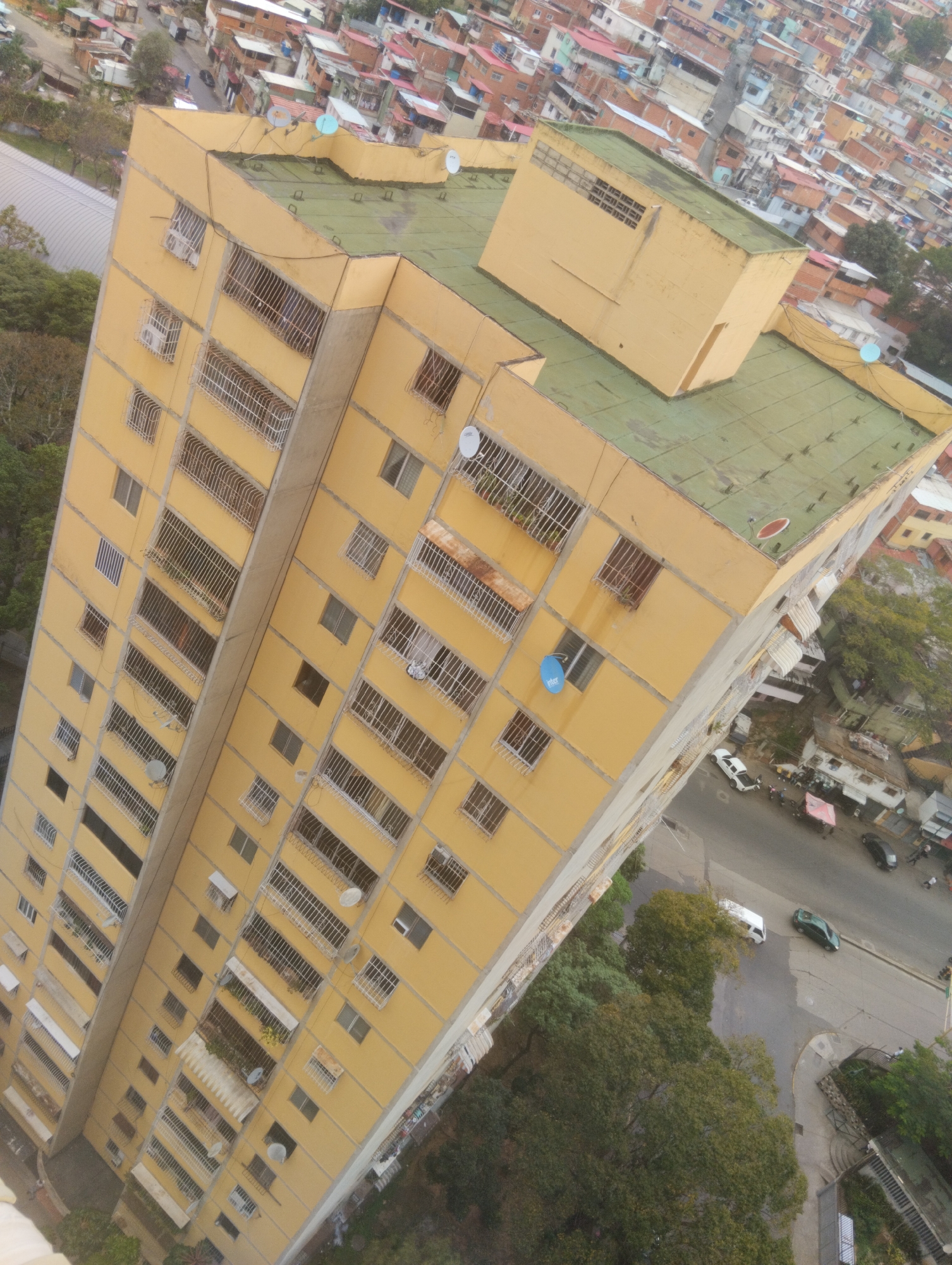 Here is a detailed description of the image:
**Scene Overview:**
The image depicts a tall, yellow apartment building in an urban setting, viewed from an elevated perspective. The building dominates the foreground and middle ground, with a sprawling cityscape of smaller buildings extending into the background.
**Building Details:**
The apartment building has a rectangular shape and is painted a bright yellow. It has numerous windows arranged in a grid-like pattern across its facade. Many of the windows have metal bars or grills. Satellite dishes are visible attached to some of the balconies and windows. The building's roof is green and has a smaller, rectangular structure on top.
**Surrounding Environment:**
The surrounding cityscape is dense and cluttered with a mix of residential and commercial buildings. Many of the buildings in the background appear to be smaller, low-rise structures with corrugated metal roofs, painted in various colors. Streets and sidewalks weave between the buildings, with trees lining some of the roads. Several parked cars are visible on the streets below.
**Composition and Lighting:**
The image has a somewhat tilted perspective, likely taken from a high vantage point within another building. The lighting appears to be natural daylight, with shadows casting on the building's facade. The sky is not visible in the image.
**People, Actions, Interactions:**
There is no indication of people or actions/interactions in the image.
**Overall Impression:**
The image gives a sense of a densely populated urban area in a developing country. The apartment building is a prominent landmark in the cityscape.