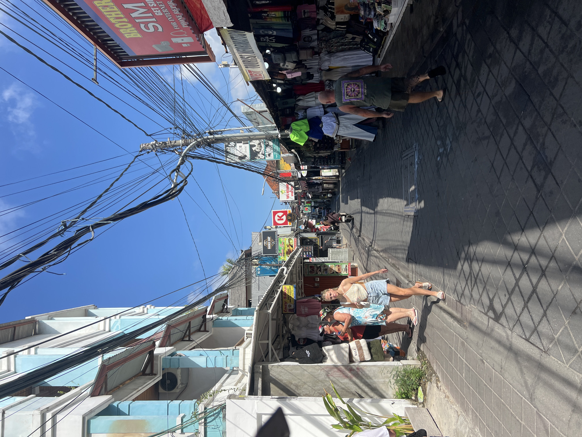 The image depicts a busy street scene in Kuta, Indonesia, characterized by narrow buildings, overhead electrical wires, and various market stalls lining the roadway. The architecture appears to be a mix of light blue and white structures with awnings extending over the sidewalks.
Several individuals are visible walking along the street. In the foreground, a woman in a white halter top and denim shorts walks away from the camera, her shadow cast on the paved ground. To her left, two other individuals, one in a red shirt and the other in a floral dress, are looking at items displayed outside a shop. Further down the street, a man wearing a green t-shirt with a purple floral design and khaki shorts walks away from the camera. In the background, a person is riding a scooter.
The market stalls display a variety of goods, including clothing, bags, and possibly souvenirs. Signs for businesses such as "BROTHER SIM," "LUCY SHOP," "BARBER SHOP," and "TURTLE RESTAURANT SPORT BAR" are visible, indicating a commercial area. The sky is clear blue with some scattered clouds. The ground is paved with interlocking square tiles, and shadows from the buildings and overhead wires are prominent. The overall impression is one of a vibrant, bustling street in a tropical location.