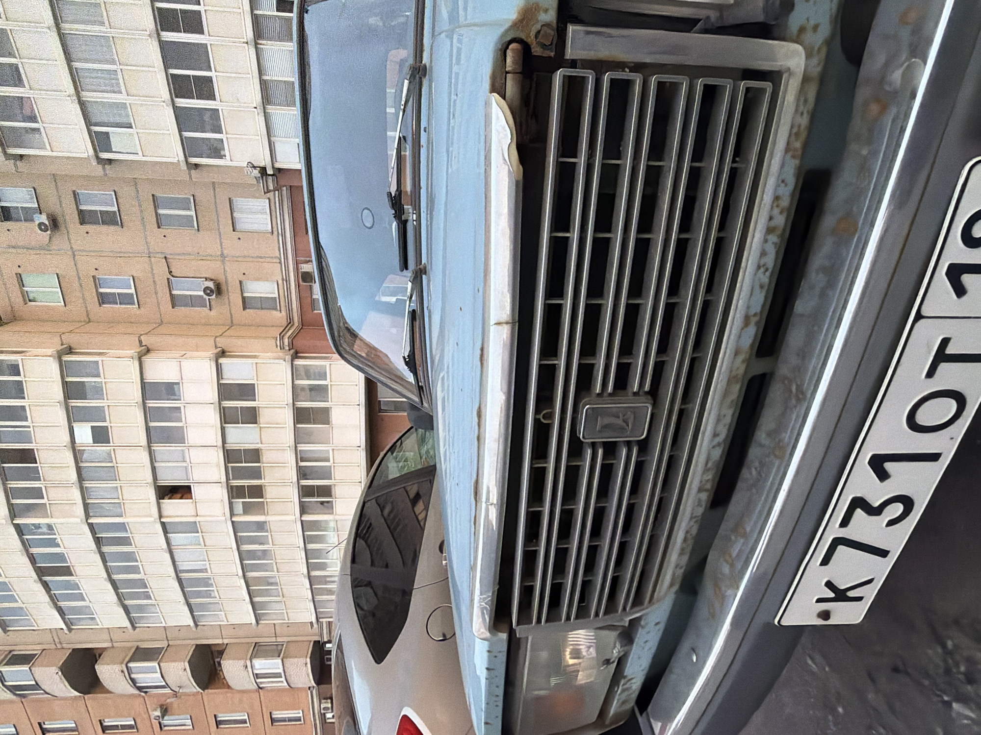 A close-up, eye-level medium shot shows the front of a light blue, slightly rusted older model car. The car's grille, made of horizontal silver bars, is prominent, with a dark rectangular emblem featuring a silver symbol in the center. A portion of the car's hood is visible above the grille, showing some rust and peeling paint. The car's license plate, a white rectangle with black characters, is mounted on the bumper. The characters read "K731OT". Behind the car, a neutral-colored building with multiple windows and balconies can be seen, suggesting an urban setting. A sliver of a silver car is visible to the left of the blue car. The overall lighting suggests an overcast day.