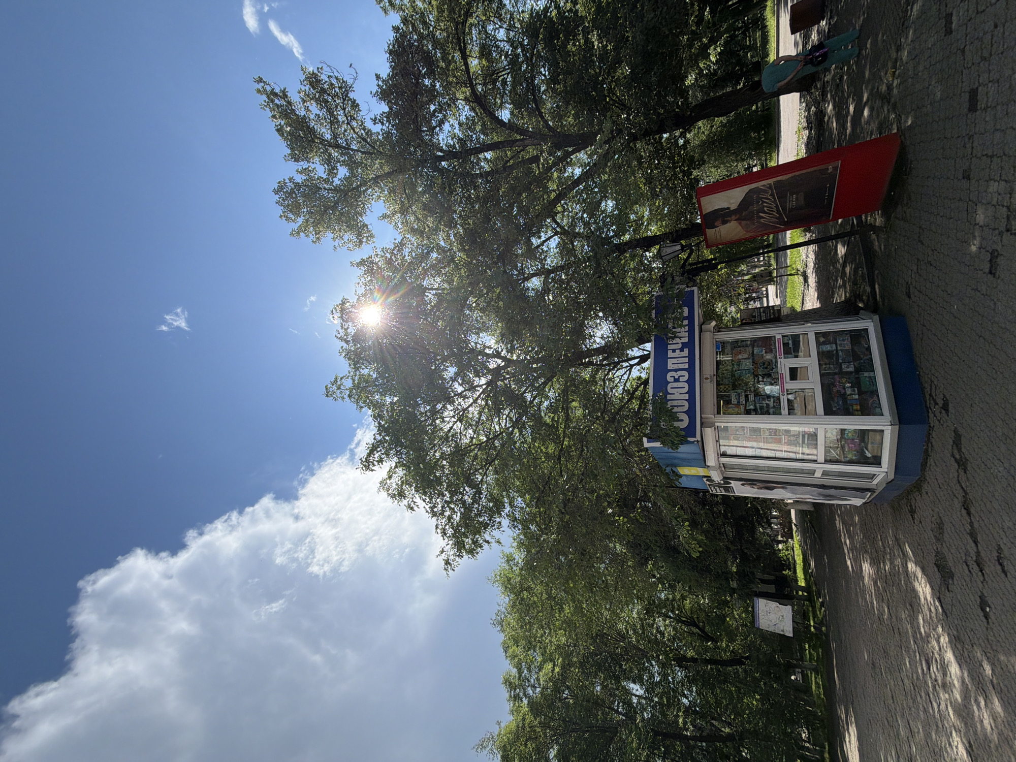 The image depicts an outdoor scene in Birobidzhan, Russia, under a bright, partly cloudy sky. A kiosk, labeled "СОЮЗПЕЧАТИ" (Soyuzpechat), which is a chain of Russian newsstands, stands prominently in the foreground. The kiosk has large glass windows displaying various items, likely newspapers, magazines, and snacks. To the right of the kiosk, a tilted red advertising stand features a poster with a man's image and Russian text. Beyond the kiosk, a pathway paved with interlocking stones is visible, shaded by large trees with lush green foliage. Sunlight filters through the leaves, creating dappled shadows on the ground. In the background, a person in a teal outfit is walking away from the camera on the right side of the frame. Another sign with a map or informational display stands to the left of the pathway. The environment appears to be a park or a public area within the city.