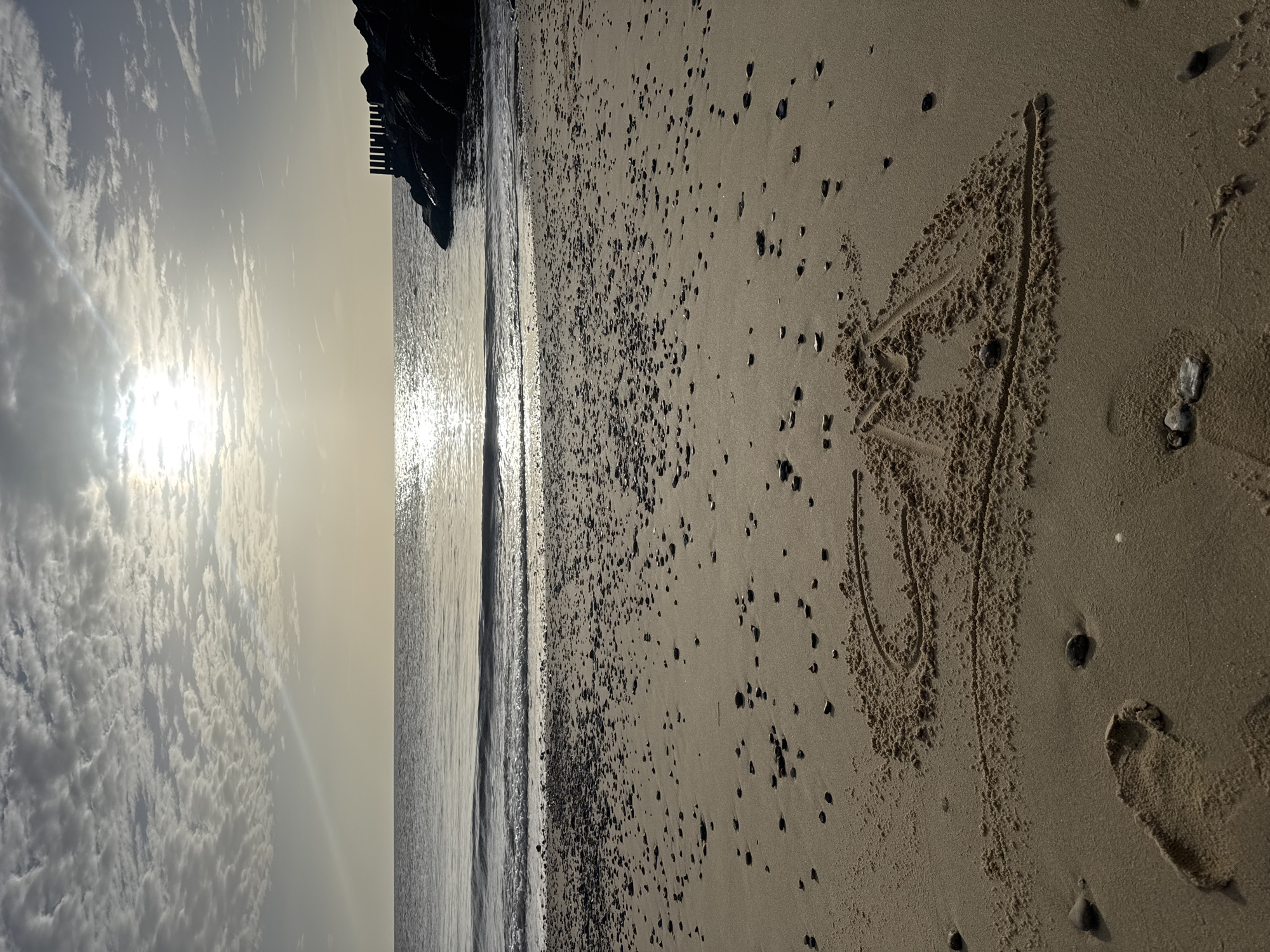The image depicts a beach scene in East Suffolk, United Kingdom. The sky is overcast with the sun obscured by clouds. The sea is calm with small waves reaching the shore. A dark, possibly rocky, structure extends into the sea on the left side of the frame. The beach is sandy, with small dark pebbles scattered across the surface. A heart is drawn in the sand, and a single footprint is visible nearby.
