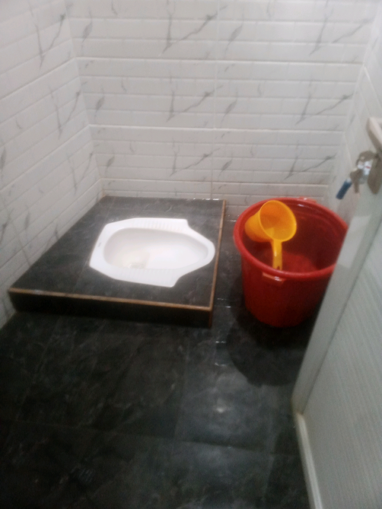 A squat toilet is installed on a raised platform covered with dark gray marble-patterned tiles. The toilet is white and has a distinctive oval shape. Next to the platform, a red plastic bucket with two yellow ladles inside is placed on the floor, which is also covered with the same dark gray tiles. The walls are tiled with white rectangular tiles that have a subtle gray marble pattern. The scene appears to be a bathroom in a residential or public facility in Randuagung, Indonesia.
