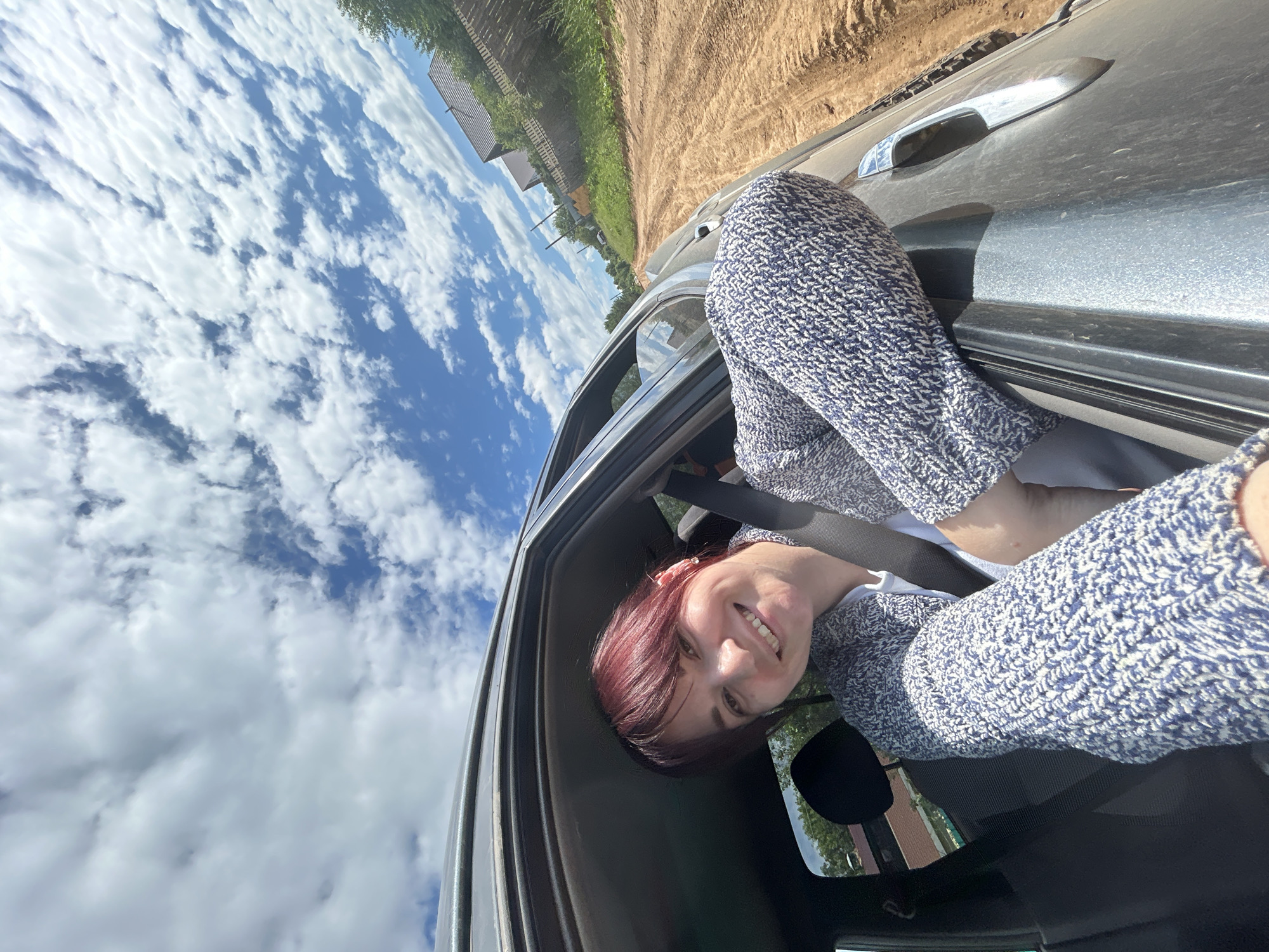 A woman with short, dark red hair is leaning out of a car window, smiling at the camera. She is wearing a blue and white patterned sweater. The car appears to be on a dirt road, with a field of green grass and some buildings to the left. The sky is blue with fluffy white clouds, suggesting it is daytime and the weather is fair. The woman's legs are also visible, tucked inside the car. The scene is set in a rural or semi-rural area, with a sense of openness and natural light. The overall mood is cheerful and relaxed, conveyed by the woman's smile.