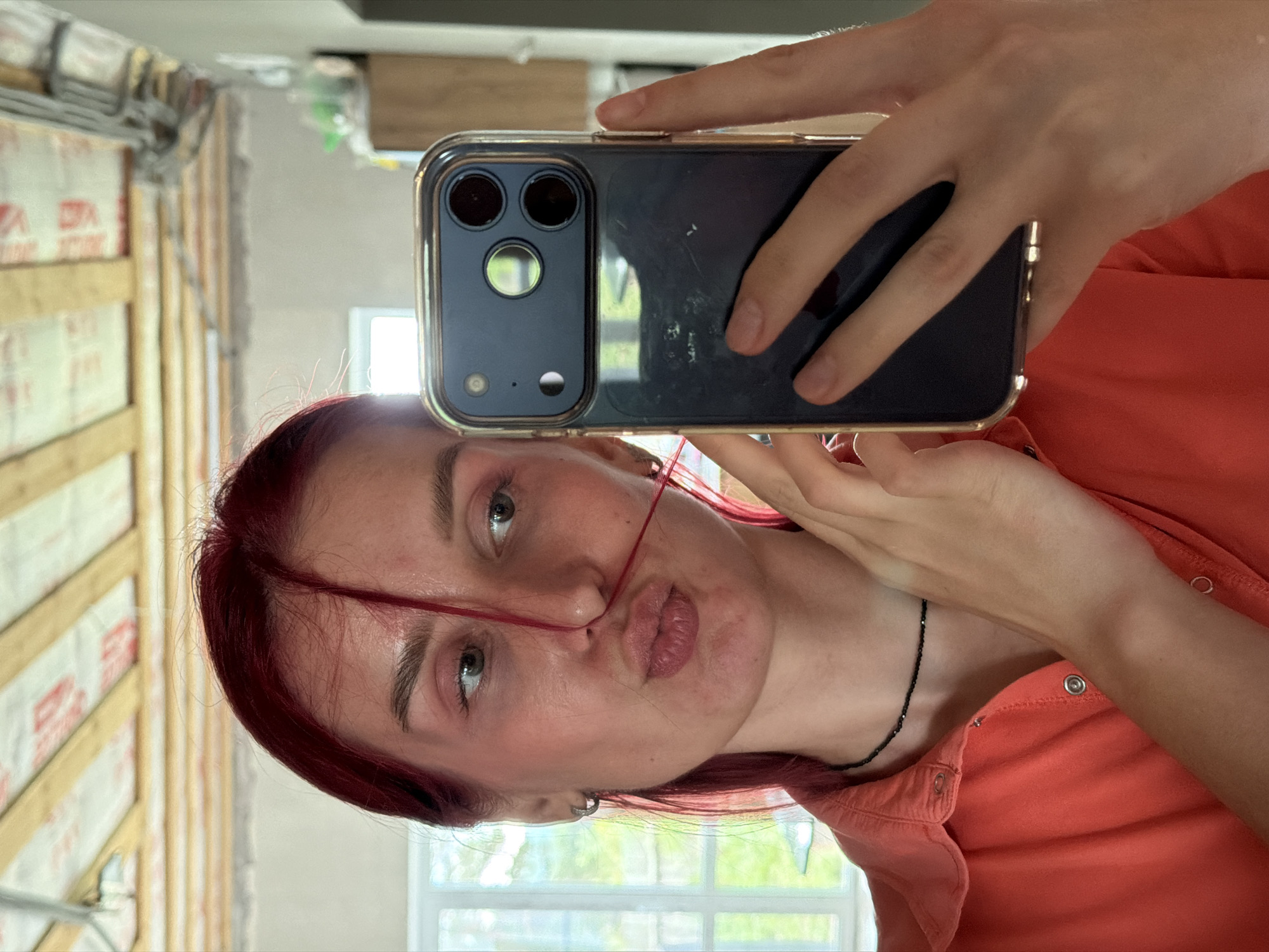 A young person with red hair is captured in a mirror selfie, making a playful "fish face" with pouty lips. They are wearing a coral-orange collared shirt with metal snaps and a thin black beaded necklace. A silver hoop earring is visible on their right ear. A strand of red hair is draped across their face.
The individual holds a dark-colored smartphone, likely an iPhone Pro model with three camera lenses, encased in a clear protective cover, which is prominently featured in the reflection.
The setting is an interior space that appears to be under construction or renovation, featuring exposed wooden wall studs or framing and white insulation material with some indistinct red printed text visible on it. Natural light streams through a window in the background, revealing greenery outside, indicating it is daytime. The image is rotated 90 degrees clockwise. The overall mood is casual and lighthearted. This scene is located in Chaikovsky, Russia.