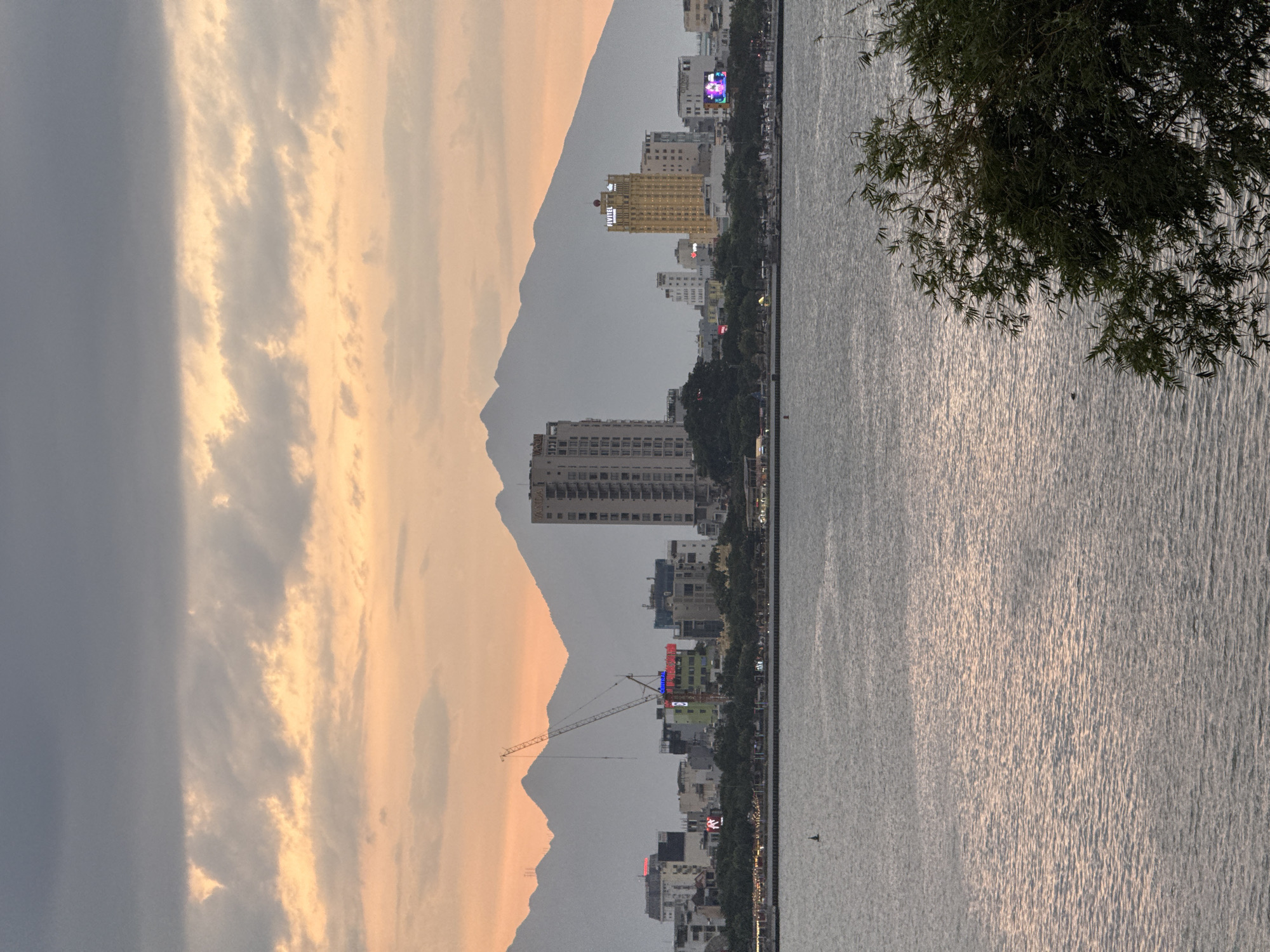 The image depicts a serene waterside cityscape at dusk in Đà Nẵng, Vietnam. The left side of the frame is dominated by a dramatic sky with soft, warm-toned clouds illuminated by the setting sun, casting a gentle glow on the imposing silhouette of a mountain range in the background. To the right of the mountains, a collection of buildings, ranging from mid-rise apartments to taller hotels, forms a bustling urban skyline. A prominent yellow building with the text "FLYITEL" is visible, along with other structures displaying illuminated signage. A construction crane stands tall amongst the buildings, hinting at ongoing development.
The right side of the image features a wide expanse of water, likely a river or bay, reflecting the muted colors of the sky and city. The water's surface is textured with ripples, creating shimmering patterns where the light catches it. A few dark, leafy branches of a tree overhang the water on the far right, adding a natural element to the scene. A small dark object, possibly a buoy or a distant boat, is visible on the water. The overall atmosphere is peaceful and tranquil, with the fading light creating a beautiful, contemplative mood. The time of day is clearly sunset, characterized by the warm lighting and the softened colors. The location is suggested by the architectural style and the presence of the mountains near a body of water.