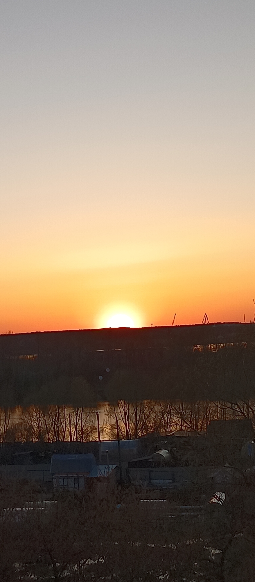 The image depicts a sunset over a cityscape of Unknown, Unknown. The sky transitions from a light grey at the top to a vibrant orange near the horizon where the sun is setting. Below the horizon, there is a dark landmass with what appears to be a river or body of water reflecting some light. Silhouetted trees line the riverbank. In the foreground, there are indistinct buildings and vegetation. In the distance, on the horizon, some cranes or industrial structures are visible. The general scene suggests an urban or suburban environment with industrial elements on the outskirts of the city.