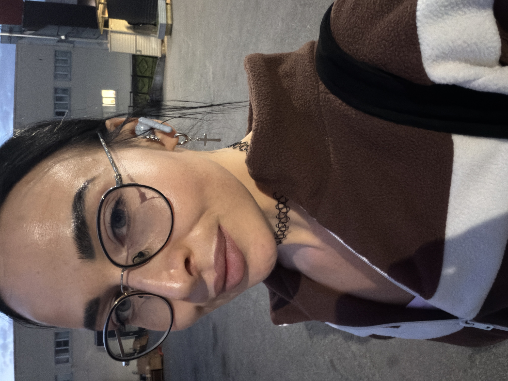 A portrait-oriented, eye-level shot captures a young person outdoors in what appears to be an urban setting in Saint Petersburg, Russia, during the evening or dusk. The subject, a woman with dark hair and a clear complexion, is looking directly at the camera with a slight, neutral expression.
She is wearing round-framed glasses, which reflect some light. An earbud is visible in her right ear, along with a silver earring shaped like a cross. Around her neck is a black choker necklace. She is dressed in a warm-looking brown fleece jacket with bold white stripes and a black accent band. A backpack strap is visible over her left shoulder.
The background consists of indistinct buildings, possibly commercial or industrial, with several windows, one of which shows a visible light, suggesting interior illumination in the darkening environment. The ground is a paved surface, likely concrete or asphalt. The overall lighting is dim, characteristic of twilight or early night. There is no visible text in the image.