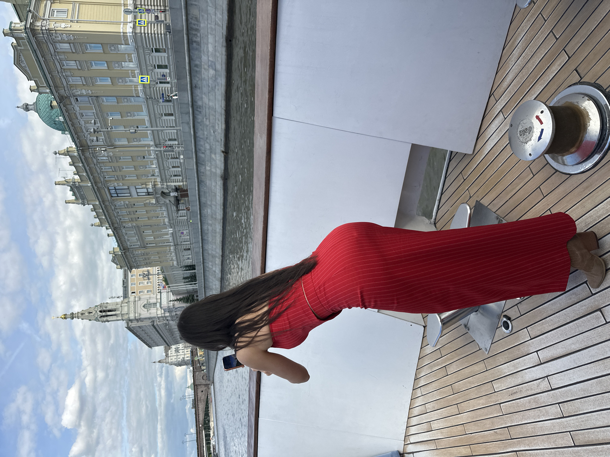 A photograph taken in Moscow, Russia, shows a woman wearing a long, red pinstripe dress and brown shoes, leaning over the side of a boat and taking a picture with her mobile phone. The boat has a wooden deck with a mechanical device on it. In the background, across the river, are several buildings, including a yellow one with a green dome and a tall white spire. The sky is partly cloudy.