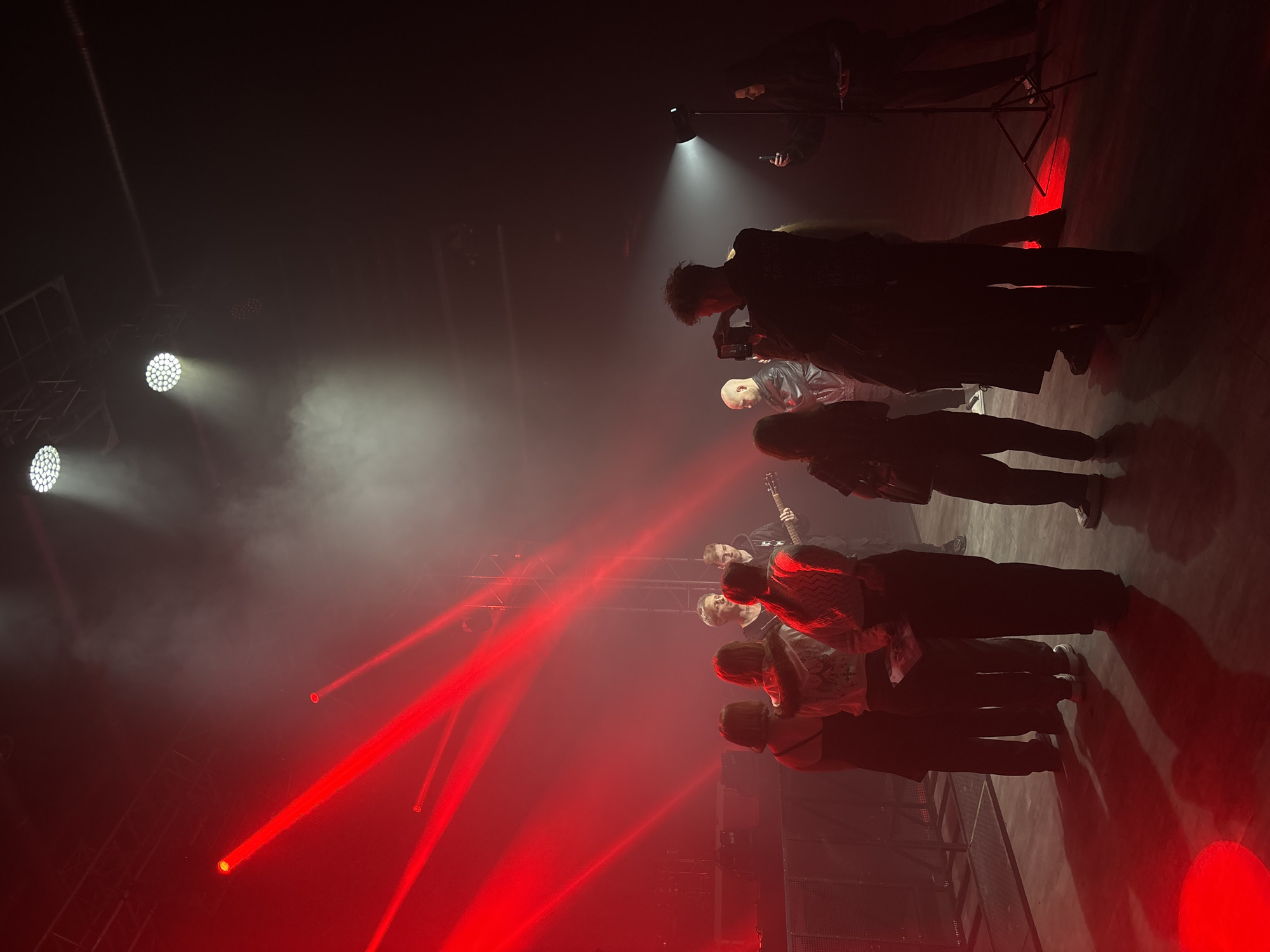 The image shows a scene inside a dark, possibly indoor concert or performance venue, possibly in Moscow, Russia. There is thick haze in the air, illuminated by both white and red stage lighting.
A group of people is standing near the foreground, appearing to watch or photograph an event. The individuals are arranged in rows. Details are obscured by shadows and lighting effects.
Further back, individuals are holding cameras, seemingly taking photos or videos of something happening. A standing lamp with a bright, downward-facing light is visible among them.
The environment is dominated by lighting and shadows, creating a dramatic and atmospheric effect. The overall impression is of an event being captured and documented.