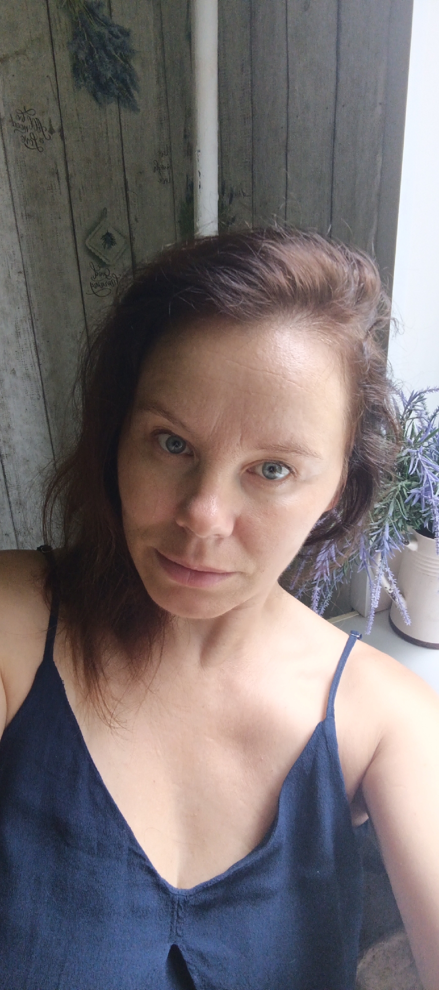 A middle-aged woman with shoulder-length auburn hair, blue eyes, and fair skin is pictured in a close-up, selfie-style shot. She is wearing a dark blue, low-cut camisole. The background consists of a distressed wooden wall with a pattern of what appears to be dried lavender branches and decorative script. To the right of the woman, a white ceramic pot holding more lavender branches is visible, along with a white vertical pipe. The lighting suggests an indoor setting with natural light coming from the right. The image was taken in Chaikovsky, Russia.