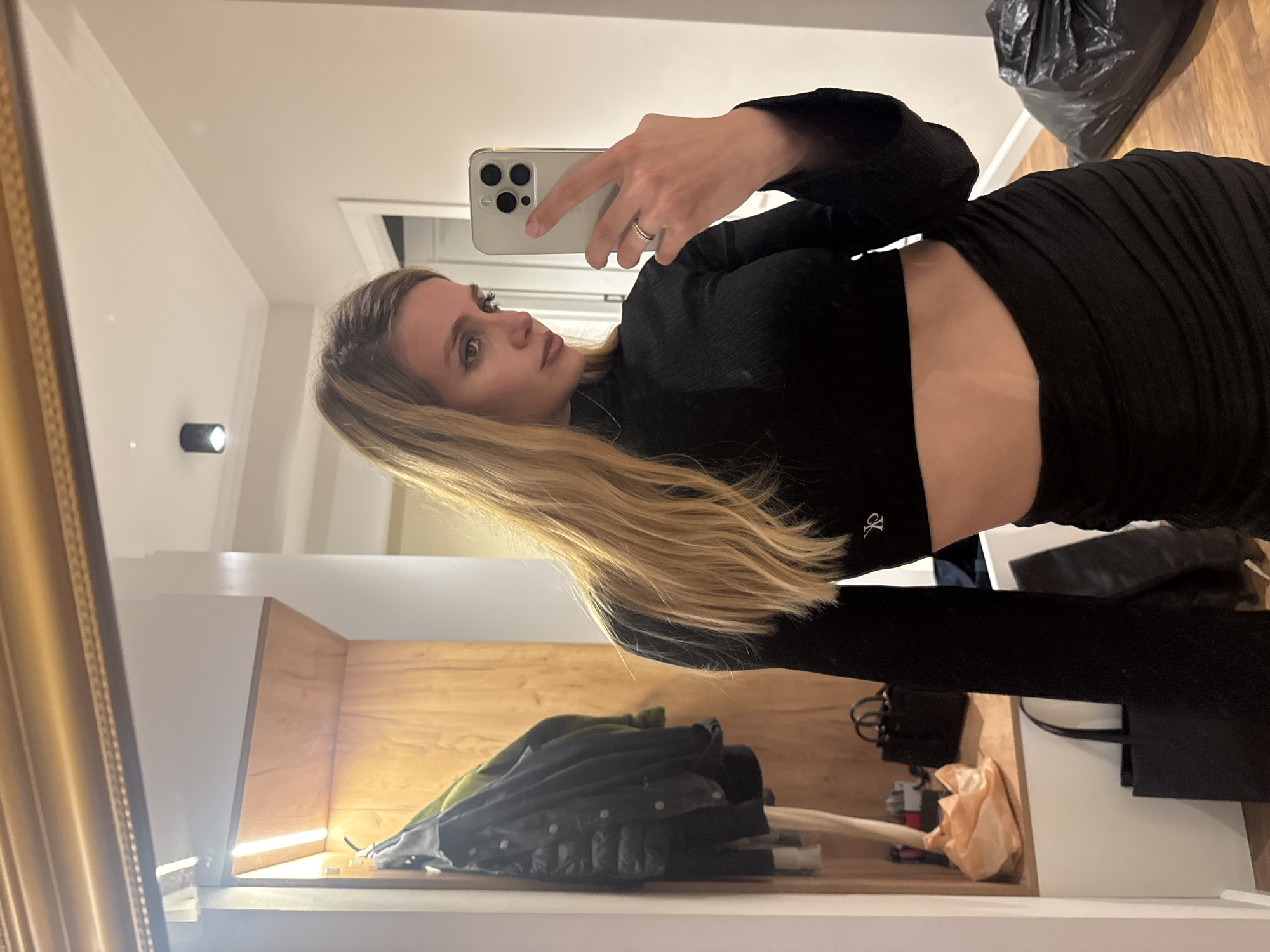 A young woman stands in front of a mirror, taking a selfie. She has long, blonde hair and wears a black, long-sleeved crop top and a tight, black skirt. She holds a silver iPhone in her right hand. The background shows a room with a shelf, a jacket, and various objects on it. In the upper-right corner, there is a black plastic bag. The reflection in the mirror suggests the scene is indoors, possibly a home. The city and specific location are indeterminate.