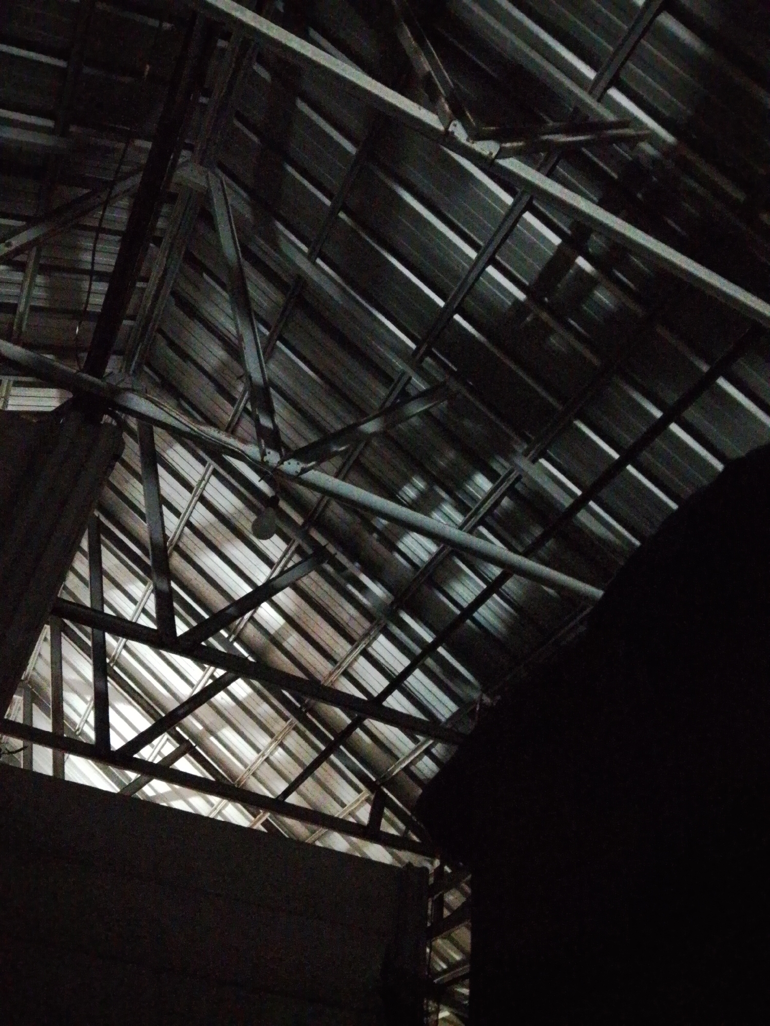 The image depicts the interior of a building in Bukit Ketok, Indonesia. The focus is on the roof structure, which consists of corrugated metal sheeting supported by a framework of metal beams. The lighting is dim, creating high contrast between the bright gaps in the roof and the darker areas of the structure. The perspective is angled upwards, providing a view directly at the roof. A large, dark shape obscures the right side of the view.