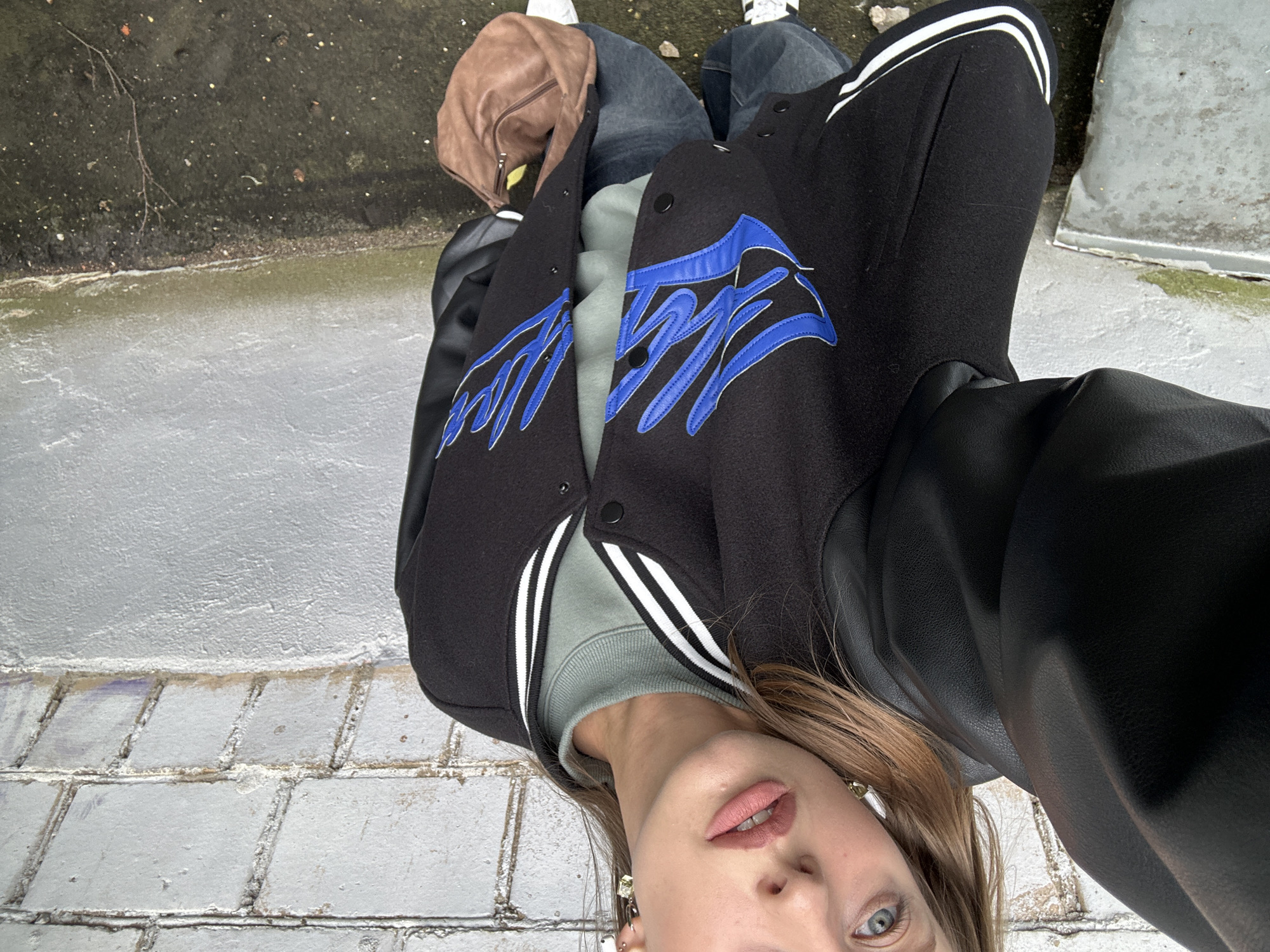 The image shows a person from the chest up, lying on their back. They are wearing a black varsity jacket with blue embroidered lettering on the front and white stripes on the sleeves. Underneath the jacket, they are wearing a light green sweatshirt. The person has long, light brown hair and is wearing earrings. Their lips are painted a matte pink color.
The background appears to be an outdoor setting with a textured grey surface, possibly concrete or pavement, and some green, murky water. There are also some small twigs and debris visible. The lighting suggests it could be daytime, but the overall atmosphere is somewhat overcast or muted. The person's expression is neutral, and the scene is static, with no clear activity taking place beyond the person being positioned in this way. The location is identified as Khimki, Russia.