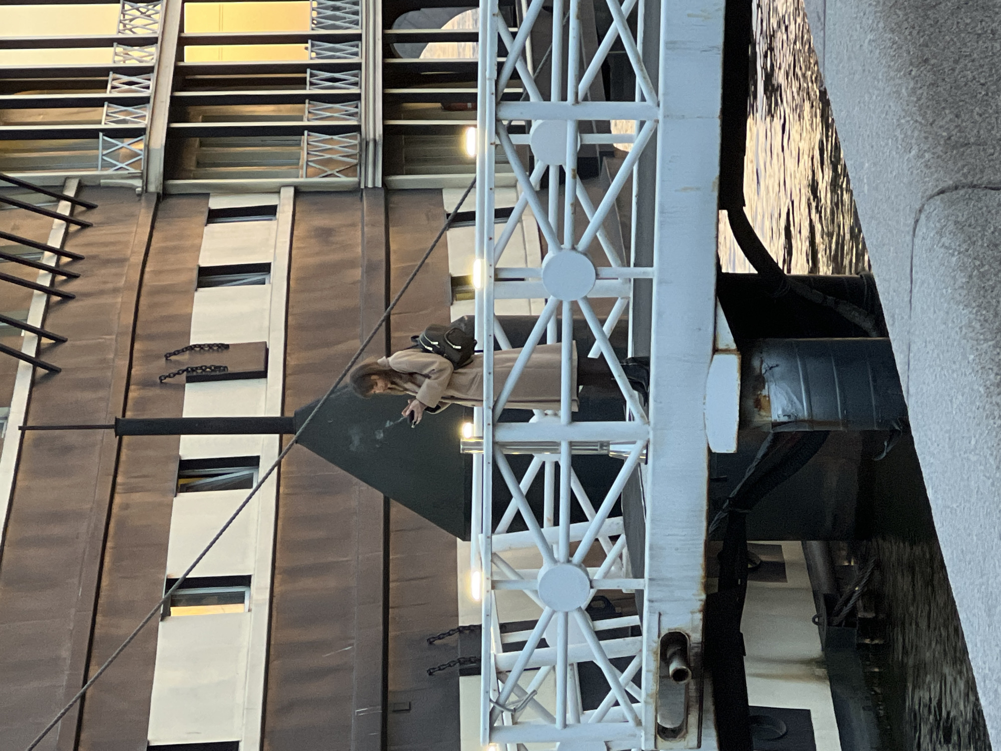 A woman stands on a bridge in Saint Petersburg, Russia, smoking a cigarette. She wears a light brown coat and a dark backpack. The bridge has a white metal framework and overlooks a canal. The building to the left of the woman has a repetitive pattern of dark vertical panels and lighter panels with windows.