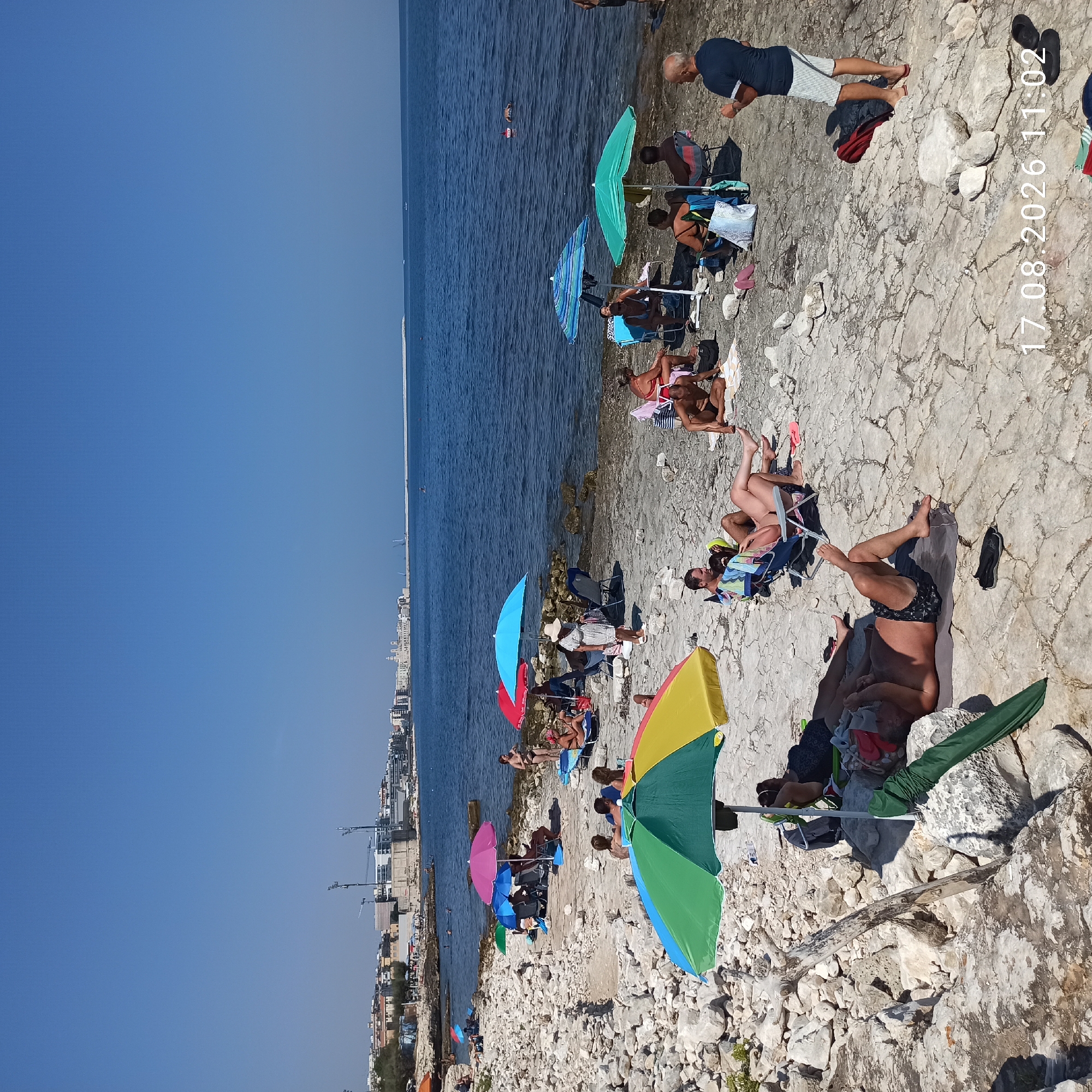 A wide-angle, eye-level shot captures a rocky beach scene in Molfetta, Italy, on August 17, 2026, at 11:02 AM. The bright sun casts sharp shadows, indicating clear weather under a deep blue sky. Several people are present, engaged in typical beach activities.
In the foreground, a person lies on a mat under a colorful beach umbrella, their legs extended. To the right, a man in a dark blue t-shirt and striped shorts stands, facing left, holding something in his hand. Scattered on the rocky ground are various beach accessories like towels, bags, and footwear.
Further back, more beachgoers are visible. Some are relaxing on lounge chairs under umbrellas of various colors, including pink, blue, and green. Others are seated directly on the rocks. In the water, a few individuals are swimming or wading, while a small boat is visible in the distance.
The background reveals the cityscape of Molfetta, with buildings lining the coast. Construction cranes and unfinished structures suggest ongoing development. The sea stretches out to the horizon, a calm, deep blue. The beach itself is characterized by large, irregular white rocks, with sparse vegetation in some areas. The overall impression is a busy, sunny day at the seaside.