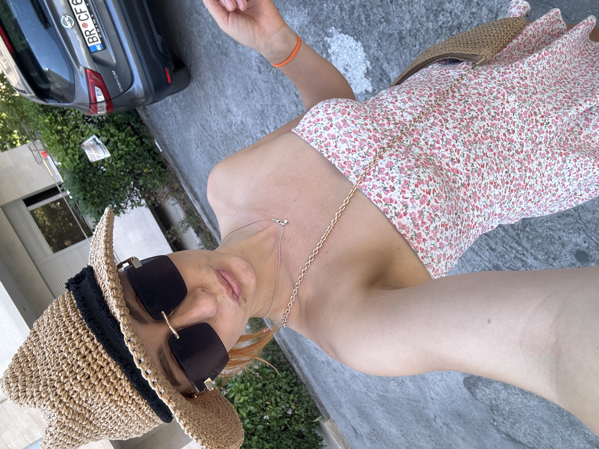 In Budva, Montenegro, a woman with auburn hair, wearing a straw hat and dark sunglasses, poses for a selfie. She is dressed in a strapless floral print dress and a gold chain necklace. An orange bracelet is visible on her right wrist. The background shows a grey asphalt surface, a green hedge, and a dark grey Nissan Qashqai with a license plate reading "BR CF 68". A sign with "ZAFFIR PARKING" is visible on the hedge.