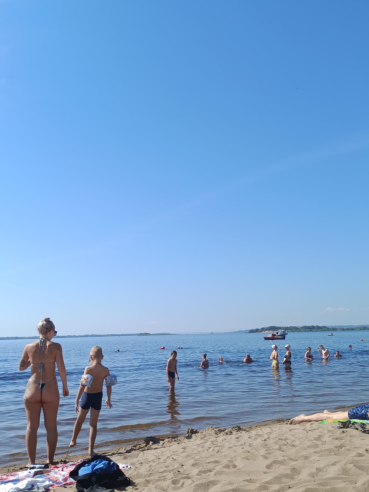 The media file depicts a sunny day at a beach in Saratov, Russia. The scene is set on a sandy shore with clear blue water stretching out to a distant horizon lined with trees. The sky is a vibrant, cloudless blue, suggesting warm, pleasant weather.
Several people are visible, both on the beach and in the water, enjoying a recreational day. In the foreground, a woman in a bikini stands on the sand, facing away from the camera, looking out at the water. Next to her, a young boy also stands at the water's edge, wearing dark swim trunks and arm floaties. Further into the water, a group of people are wading and swimming. Among them, a young boy in dark swim trunks stands in the water.
In the distance, on the water, a red and white boat is visible, and another smaller boat with people in it can be seen further to the right. The overall atmosphere appears relaxed and leisurely, with individuals enjoying the summer weather and the water. The time of day is likely midday or early afternoon, given the bright sunlight and the clear sky. No text is visible in the media file.