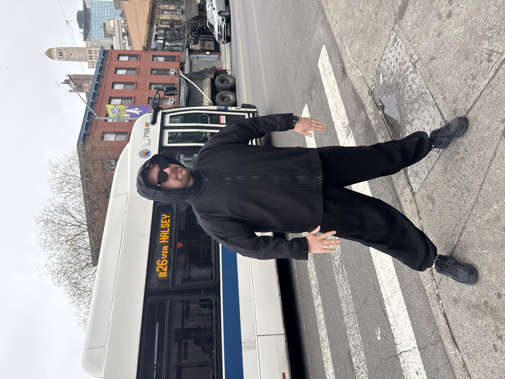 A person is standing on a pedestrian crossing next to a B26 bus. The bus reads "B26 via Halsey." The person is wearing black clothing, a hooded jacket, sunglasses, and black shoes. In the background, there are buildings and a tower. The photograph appears to have been taken in New York City, United States of America.
