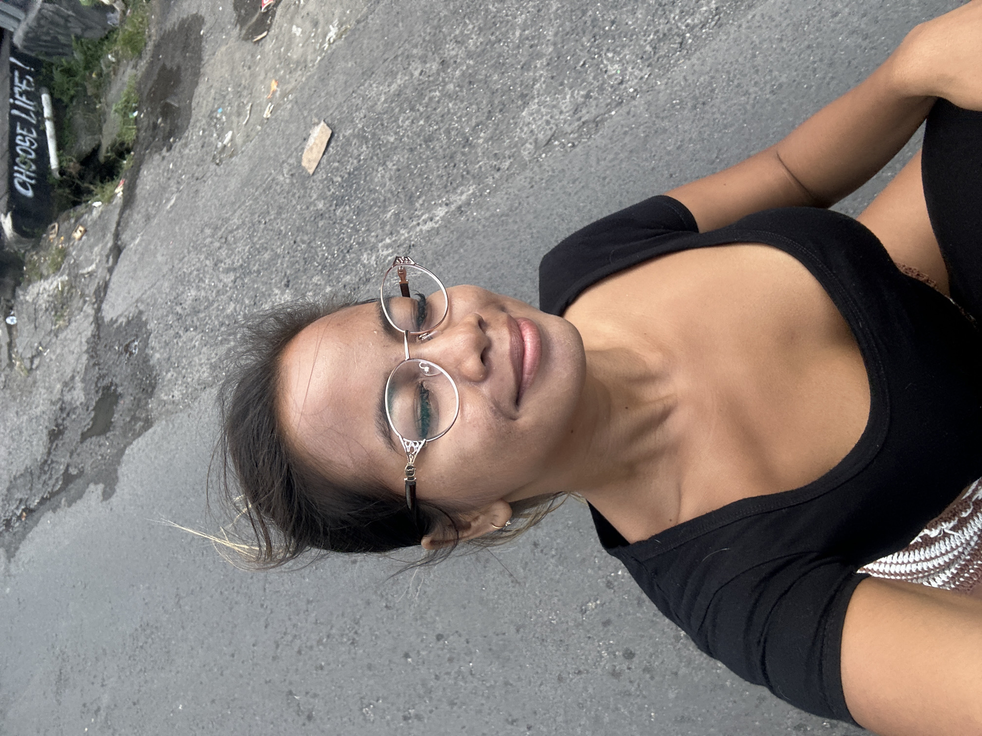 A medium shot captures a young woman from the chest up, lying on her back on an asphalt surface. She is wearing a black tank top with a low neckline and a beaded or woven detail visible at her hip. The woman is also wearing round, delicate-framed glasses. Her eyes are closed, and her lips are slightly parted, suggesting a relaxed or peaceful expression. Her dark hair is loose around her head, with a few strands catching the light.
The background consists of the grey, textured asphalt, with some debris scattered on it, including small pieces of cardboard and what appears to be litter. To the left of the woman, there is a patch of green vegetation and a wall with graffiti. One piece of graffiti clearly reads "CHOOSE LIFE!". The lighting suggests it is daytime, possibly overcast given the soft light and lack of strong shadows. The overall scene appears to be outdoors in an urban or semi-urban environment, possibly an alleyway or a less-maintained public space. The orientation of the image is such that the woman's head is towards the top left and her body extends towards the bottom right.