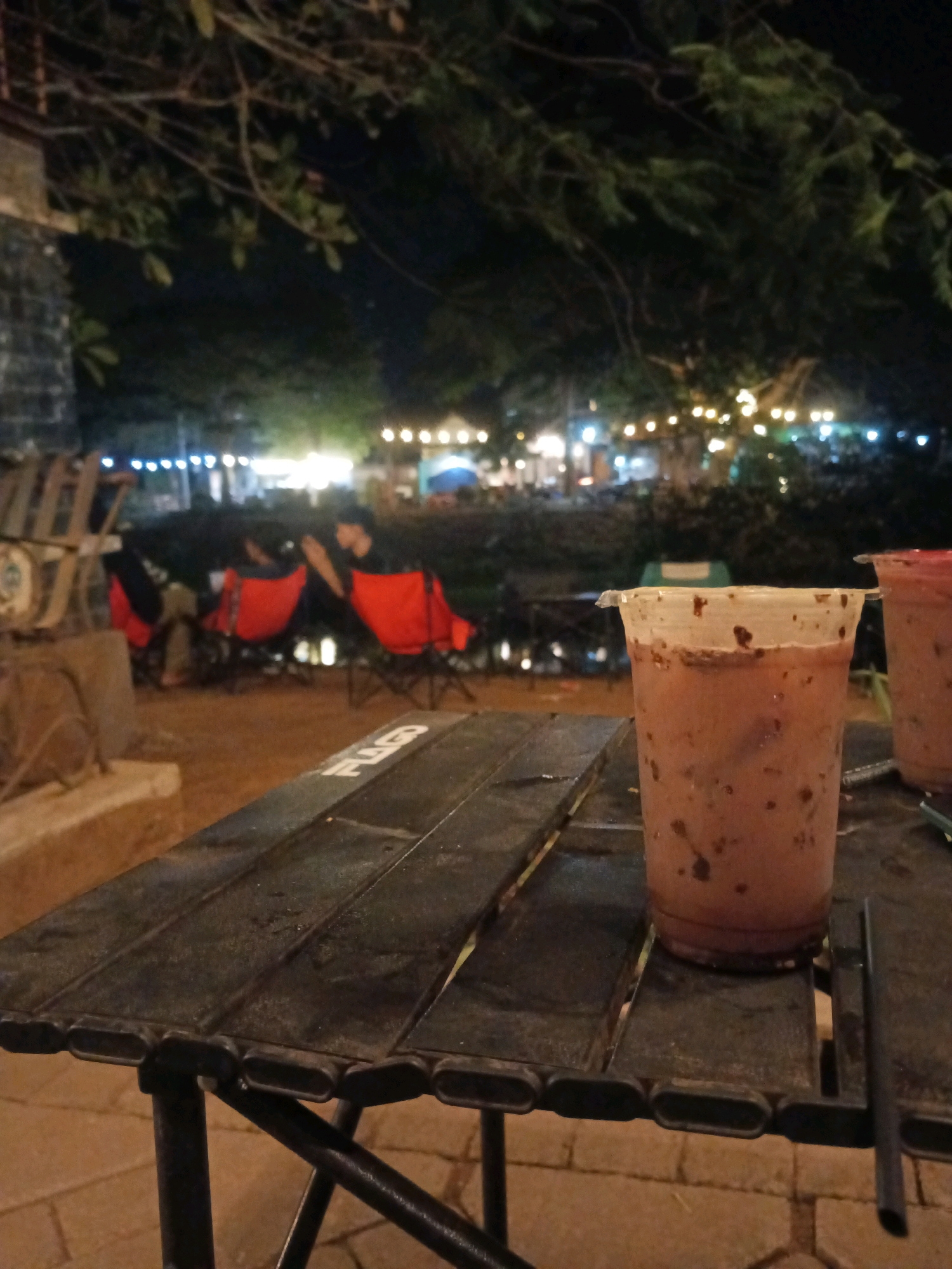 A close-up shot of a plastic cup filled with a brown, iced beverage sits on a dark, foldable table. The cup has visible flecks of chocolate or coffee grounds throughout the liquid and on the inside walls. The table's surface is made of dark, slatted material, and the legs are visible, indicating it's a portable outdoor table. In the background, out of focus, are several red camping chairs, two of which are occupied by people wearing dark clothing. One person in a chair is looking down at their hands, while another is leaning back. Beyond the chairs, a dimly lit outdoor area is visible, with string lights illuminating trees and what appears to be a gathering or dining area. The overall atmosphere suggests a relaxed evening setting, possibly at an outdoor cafe or rest stop. This scene could be located in Tulungagung, Indonesia, a region known for its various natural attractions and growing cafe culture, especially during the evenings.