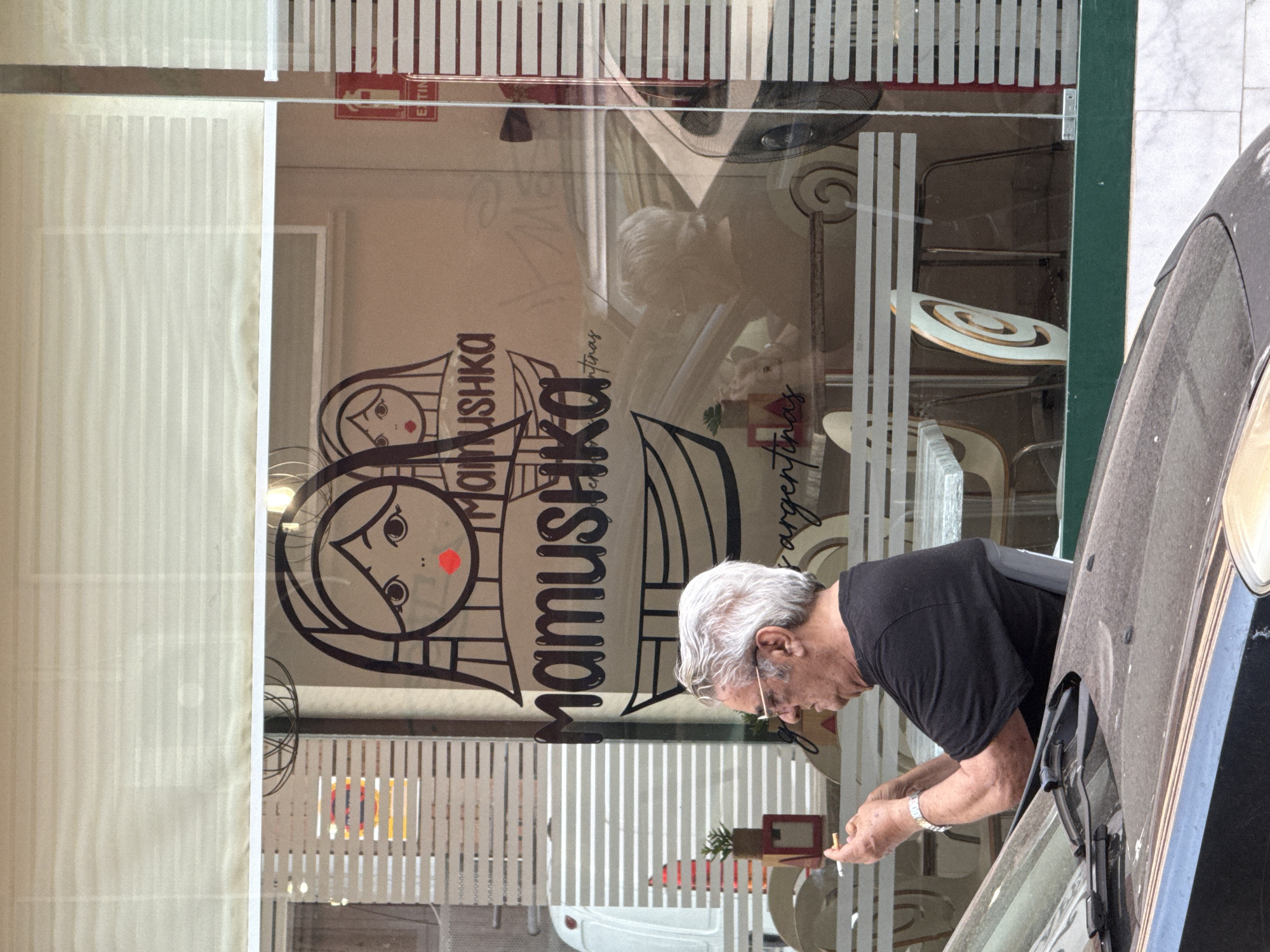 The image presents a street-level scene featuring an older man outside a commercial establishment, viewed over the hood of a dark-colored vehicle.
In the foreground, the dusty hood of a dark car, equipped with wipers, occupies the bottom left. A partial view of a headlight is visible on the bottom right.
In the midground, an older man with short, wavy grey hair and glasses is seated in profile, facing right. He wears a black t-shirt and a silver watch on his left wrist. His right hand holds a cigarette between the thumb and forefinger. The man is positioned outside a building with a large glass storefront.
The glass storefront displays a prominent black graphic logo. This logo features a stylized face of a woman with red lips, resembling a matryoshka doll, with a smaller, similar face partially visible behind it. Below this illustration, the word "Mamushka" is written in a stylized font, followed by "argentinas" and a boat-like symbol. The window also shows a series of horizontal frosted or etched lines across its lower portion.
Through the clear sections of the window, parts of the interior of the establishment are visible. These include tables with light-colored tops, chairs with distinctive white and spiraling light brown backrests, and a small potted plant in a red frame on a table near the window. Reflections on the glass show elements of the exterior environment, such as a white van and a fire extinguisher sign (partially visible as "EXTIN") inside the building. Above the storefront, beige horizontal blinds are partially lowered.