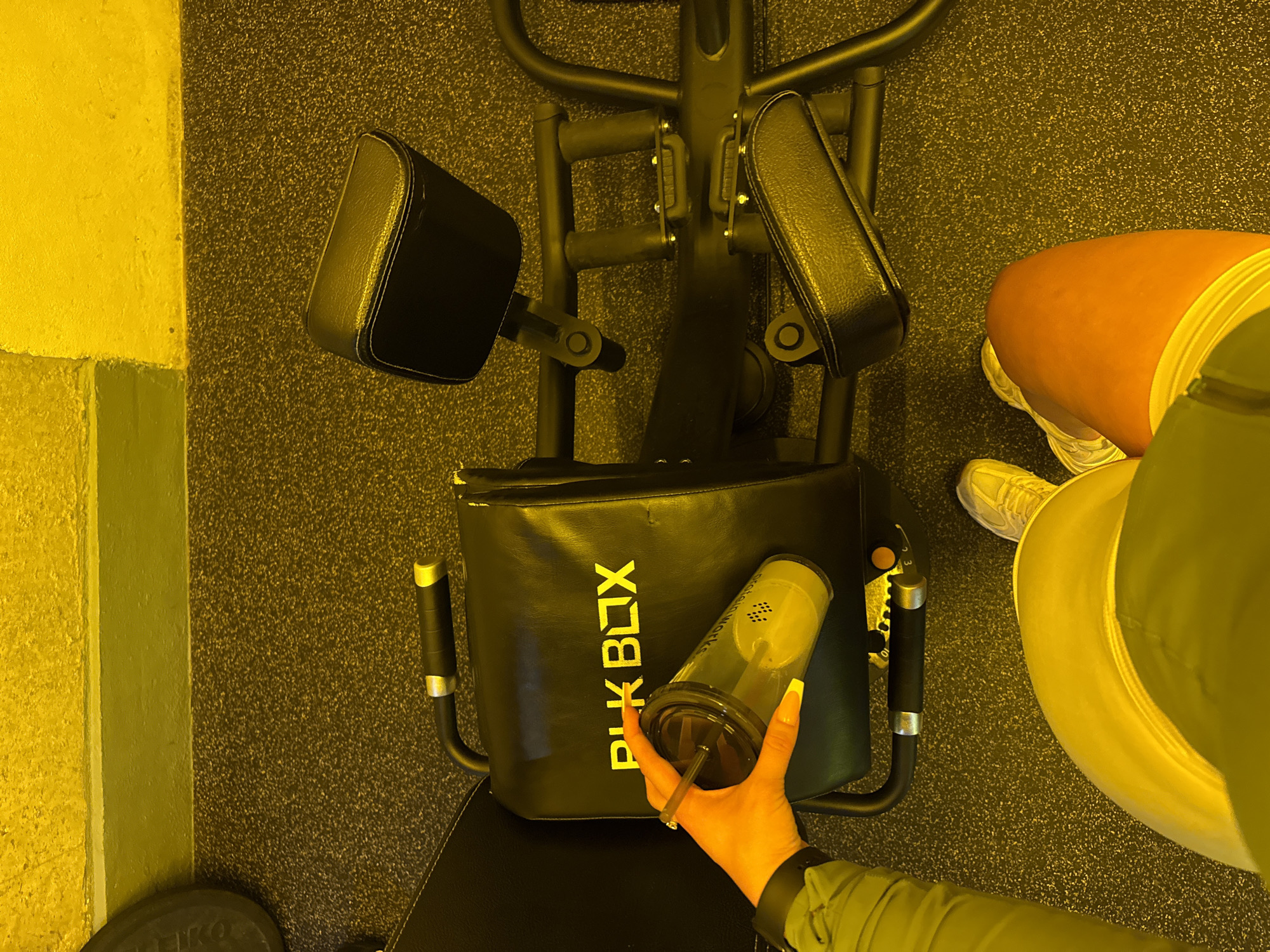 The image depicts a gym environment, with a weight plate visible on the left and gym flooring. In the center, a piece of gym equipment with black padded seating is present. The brand "BLK BOX" is visible in yellow lettering on the equipment. A person's hand, adorned with a ring and long, bright yellow-tipped fingernails, is holding a transparent cup with a dark lid and straw. The cup contains a light-colored liquid. The person is wearing a green jacket and yellow shorts, with yellow sneakers visible at the bottom of the frame. The lighting in the image casts a strong yellow hue over the entire scene. No specific location in London, United Kingdom, can be determined from the visual information.