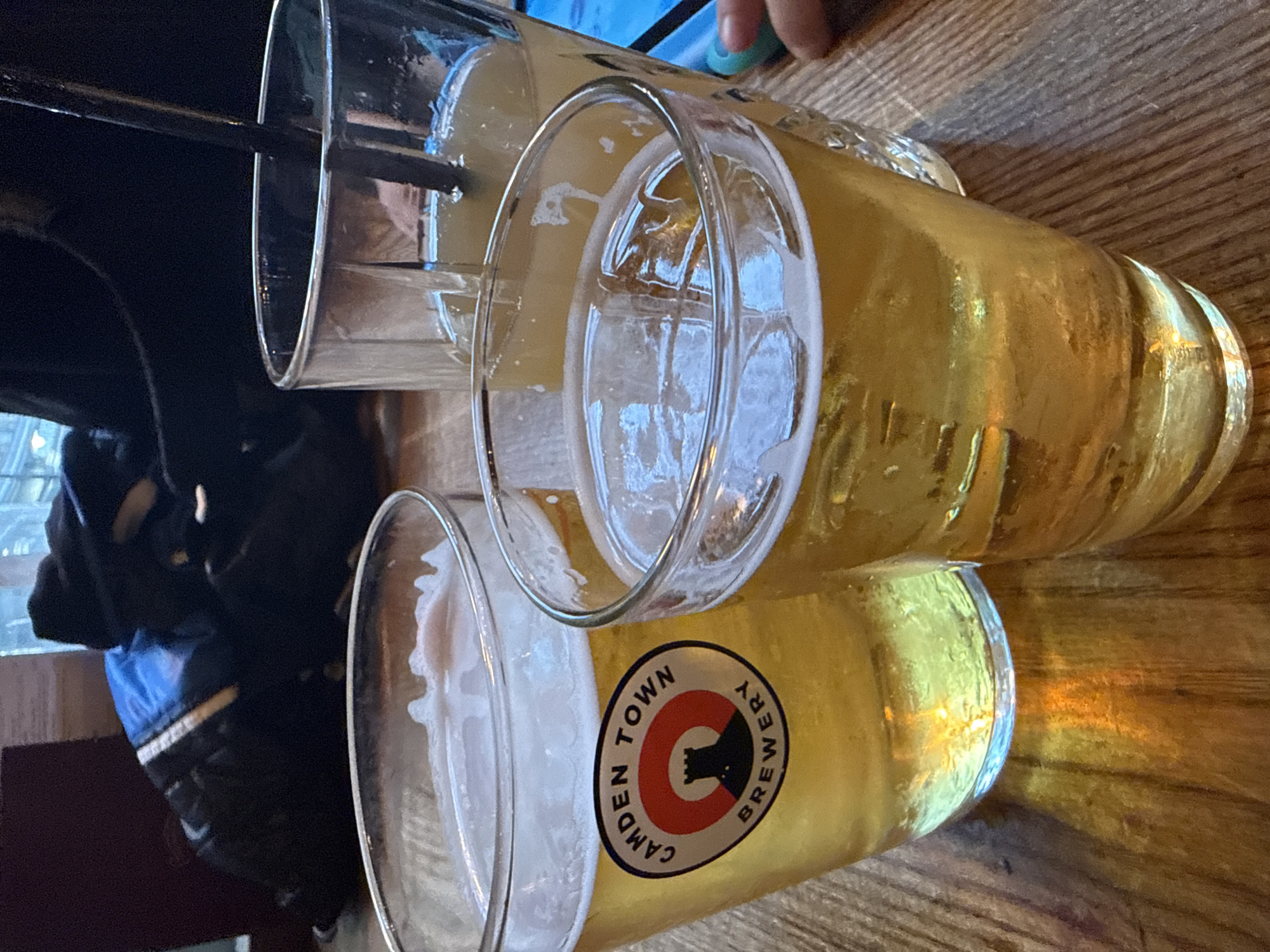 The image shows three pint glasses of beer on a wooden table. One glass has a black straw in it. The glass closest to the viewer displays a Camden Town Brewery logo. The background includes a blurred view of what appears to be an interior space, with a partial view of a person wearing a blue jacket.