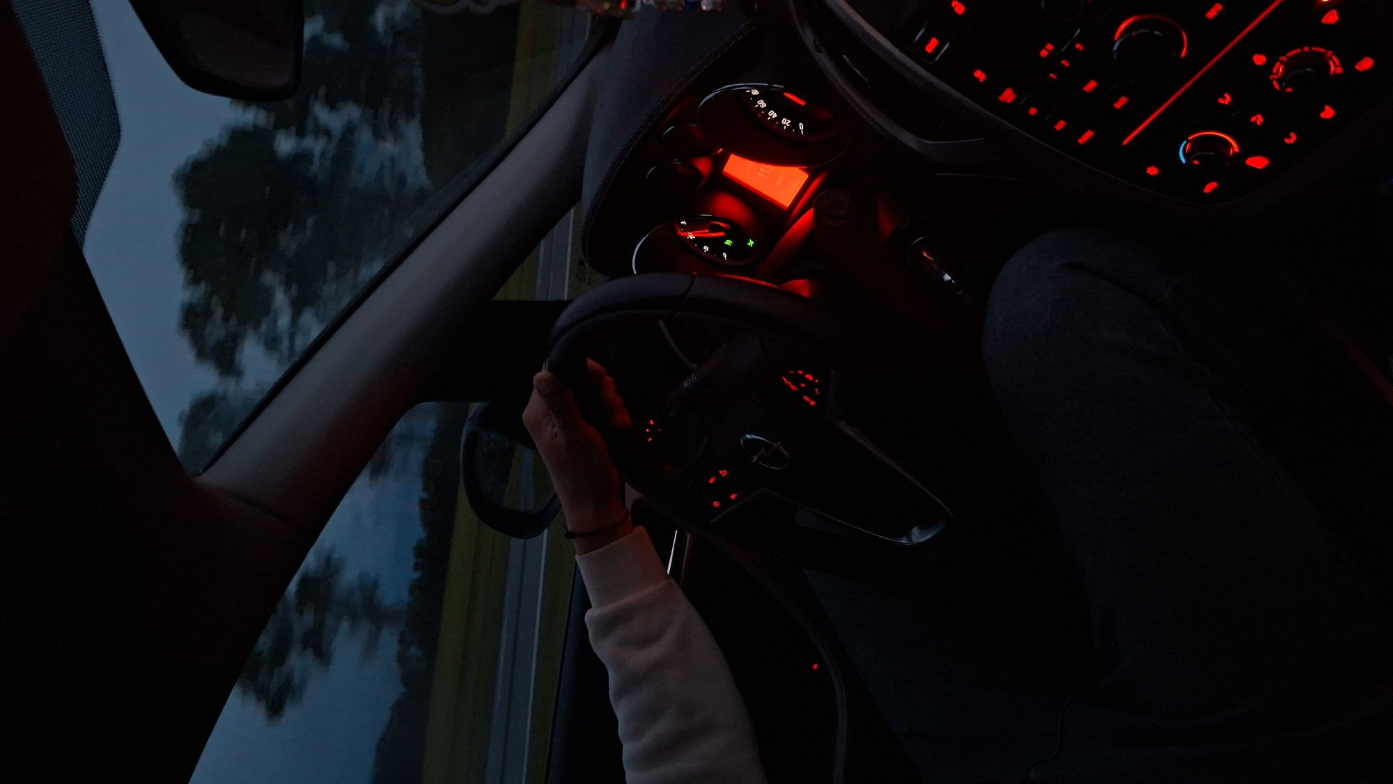 The image depicts the interior of a car at night in Самахвалавіцкі сельскі Савет, Belarus. The dashboard and steering wheel are visible, illuminated by red lights. A person wearing a white sweater is holding the steering wheel. The outside environment is visible through the car window.