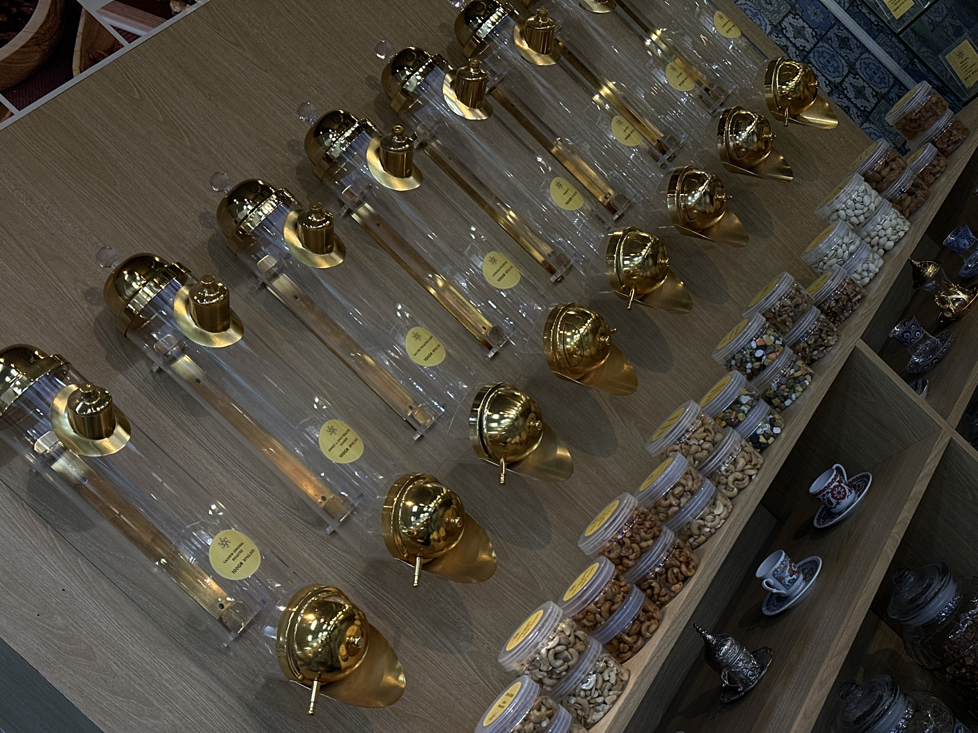 The image captures a view of a nut dispensing system in a store, likely in North Kuta, Indonesia, given the product labels with Indonesian currency and text. The system consists of clear plastic tubes with ornate, brass-colored dispensing mechanisms. Each tube is labeled with information about the contents, including varieties like "Cashew Original Roasted," "Sweet & Spicy Cashew," and "Salted Roasted." The weight of each product is marked as "100GR" (100 grams), along with a price "RP45.000," presumably referring to the Indonesian Rupiah currency. Next to the dispensing system, there are shelves stocked with clear plastic containers filled with various types of nuts, likely the same contents as the dispensers. The overall environment appears to be a well-lit, organized retail space, focusing on nuts or specialty food items.